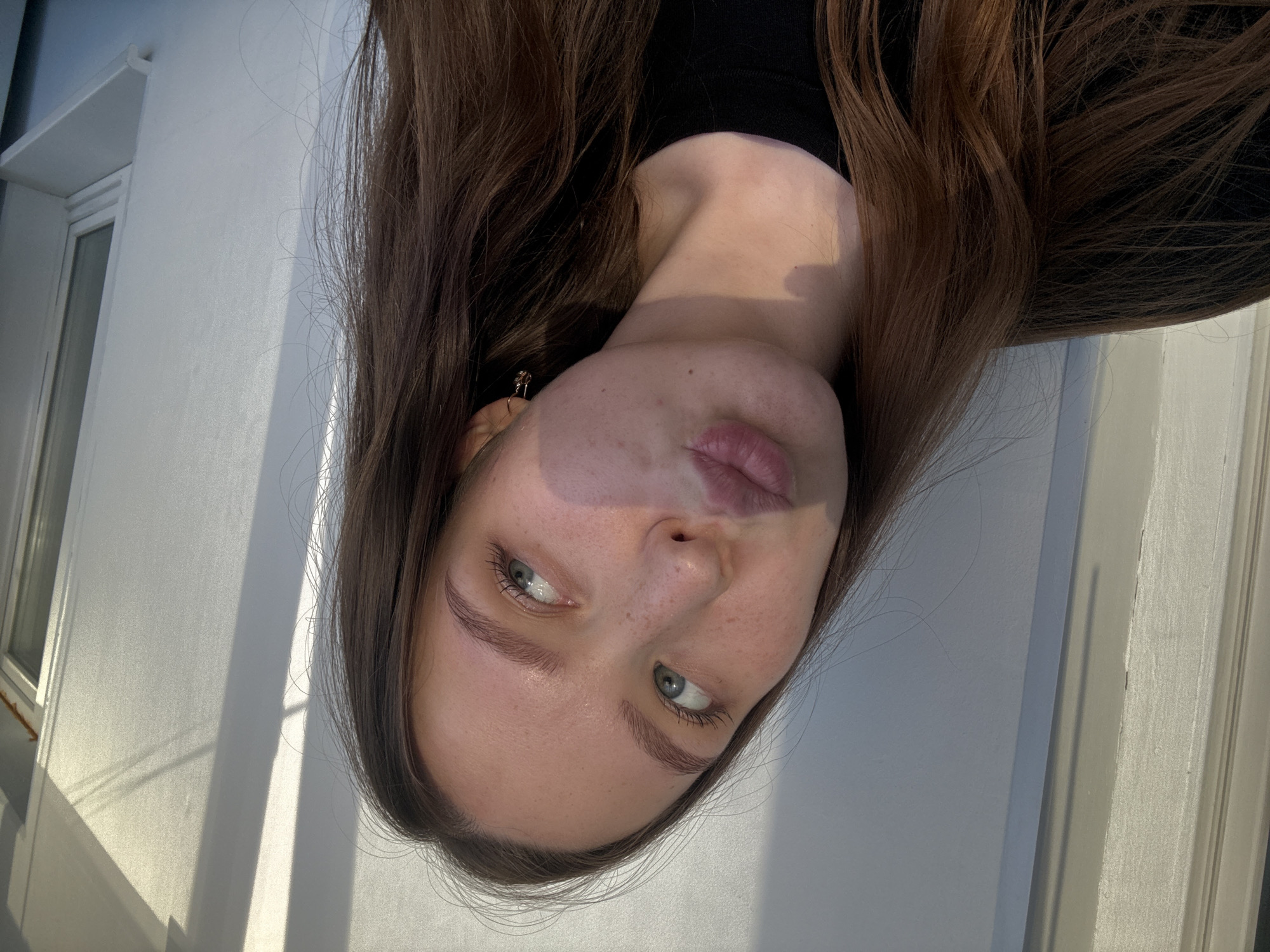 A portrait-style photograph captures a young woman with long, flowing dark brown hair. Her face is prominently featured, with her head tilted, and she looks upwards with light blue or green eyes. She has well-defined eyebrows and full, slightly parted lips, giving her expression a subtle pout or pensive look. Freckles are visible across her nose and cheeks, suggesting a natural appearance. She wears a dark-colored top, possibly black, with a small, shiny earring visible in her left ear.
The background consists of a plain, light-colored (likely white or off-white) wall. Strong, distinct shadows are cast across the wall, indicating bright, direct sunlight, suggesting it is either morning or late afternoon on a clear day. Architectural elements are visible on the left side of the frame, including a white window frame with multiple panes and a narrow ledge above it. Another vertical white structure, possibly a door frame or part of another window, appears on the right edge. The setting appears to be outdoors, possibly on a balcony or against the exterior of a building in Chaikovsky, Russia. There is no visible text in the image.