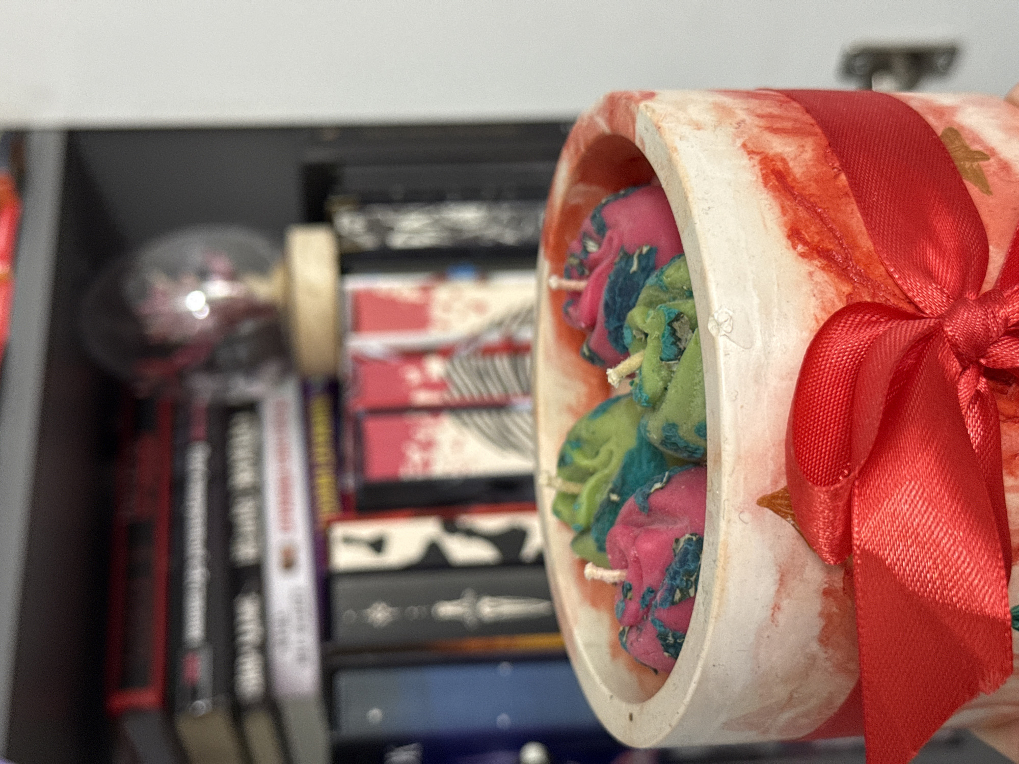 The image shows a close-up of a candle holder with several small, colorful candles inside. The holder itself appears to be made of a textured material with red and white marbling, and it is adorned with a red ribbon tied in a bow at the front. In the background, a shelf filled with books is visible. The lighting suggests an indoor setting. The specific location in Łódź, Poland, cannot be determined from the visual information provided.