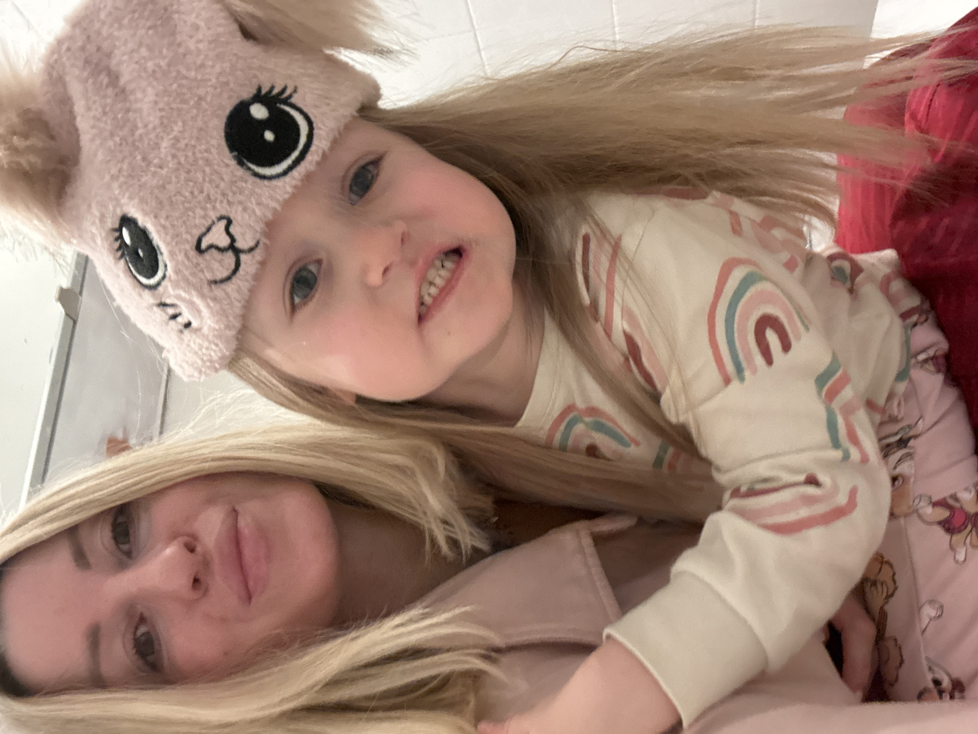 A selfie-style, indoor shot depicts a blond woman with a child, likely a mother and daughter, lying down. The child is wearing a pink hat with cartoon eyes and mouth. The child is wearing a beige long-sleeved shirt adorned with rainbow patterns. The woman's blond hair is also visible, and she is wearing a pink garment. A bed or surface with patterned bedding is visible, and a red blanket appears at the edge. The image is taken at a slightly skewed angle. The location cannot be determined from the image, but it is indicated that the photo was taken in the city of València, Spain.