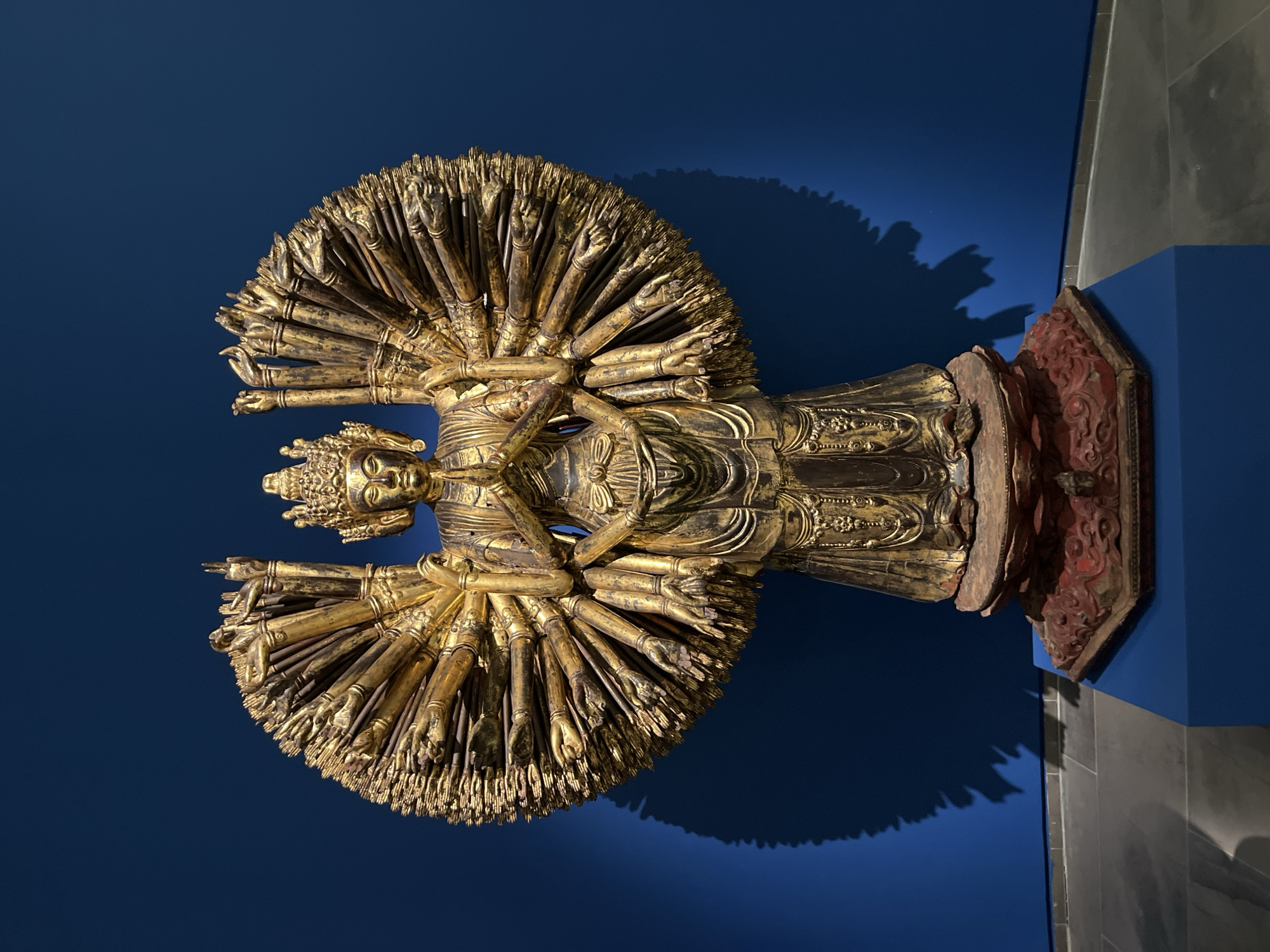 An ornate, golden statue of a multi-armed deity stands on a multi-tiered pedestal against a vibrant blue wall in a museum setting. The statue, likely depicting a Buddhist figure such as Avalokiteśvara in his Thousand-Armed form, is elaborately adorned with numerous arms fanned out behind it, each with a hand in various mudras (symbolic hand gestures). The deity wears a crown and flowing robes, detailed with intricate patterns. The pedestal is carved and painted, with the top layer appearing to be reddish-brown and the base a darker, carved wood with red accents. The statue casts a distinct, large shadow on the blue wall, emphasizing its size and the dramatic lighting of the exhibition space. The floor is a polished, grey stone. The overall impression is one of sacredness and artistic grandeur, common in museum displays of religious artifacts. The location is the Guimet Museum in Paris, France.