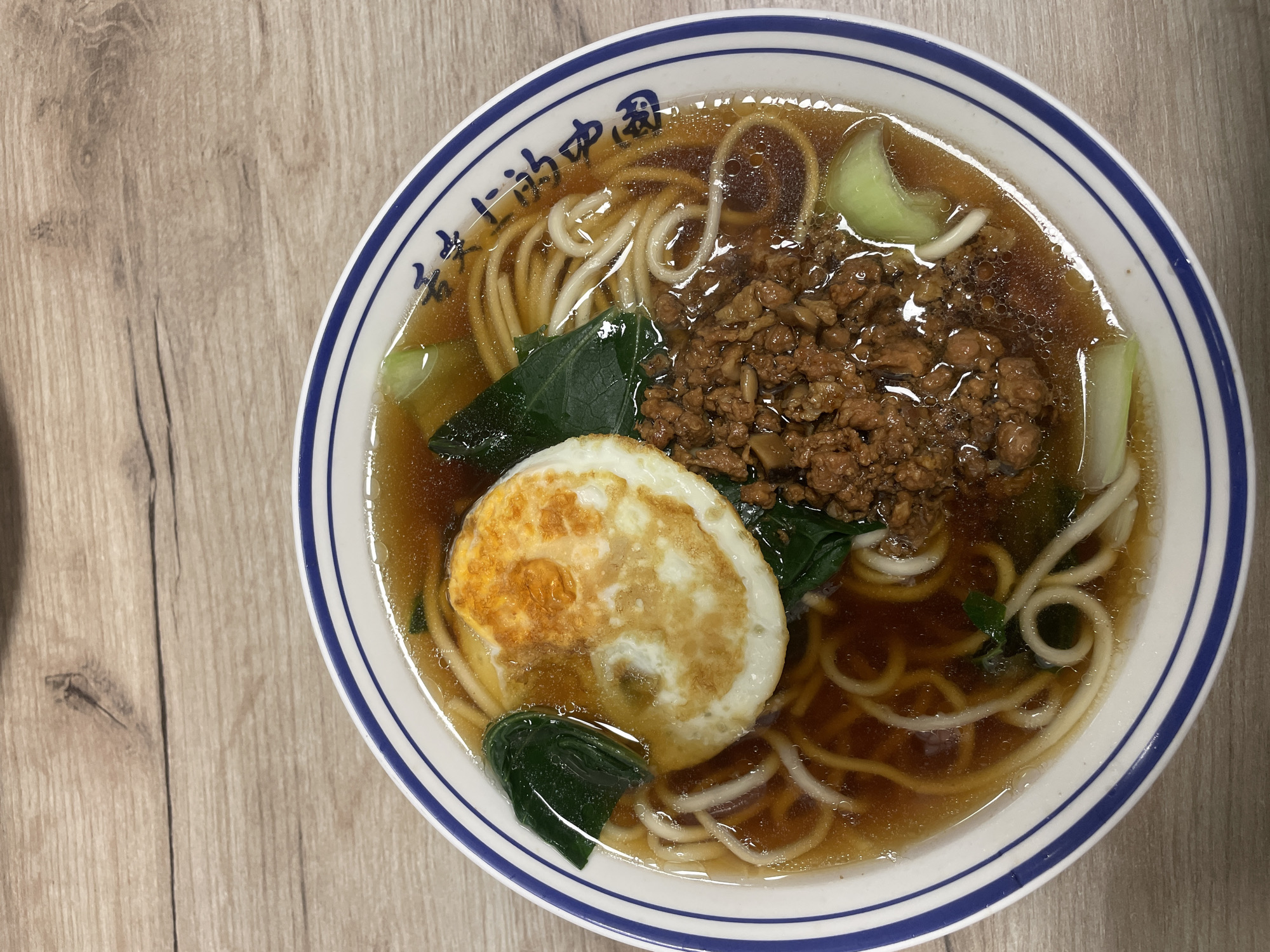A top-down shot shows a bowl of noodle soup on a light-colored wooden surface. The soup contains noodles, ground meat, leafy green vegetables, and a fried egg. The bowl is white with a blue rim and blue Chinese characters around the upper edge. The soup is a dark brown color. No people are visible. The setting is an indoor location, possibly a dining table. No specific information indicates that this photo was taken in Belgrade, Serbia.