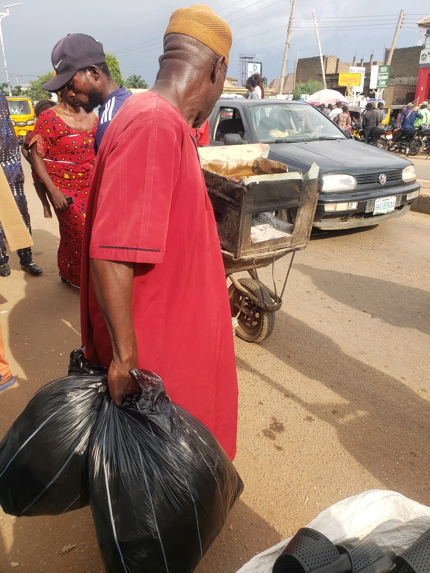 In Sabon Tasha, Nigeria, a man in a red robe and yellow cap carries two large black trash bags. He stands on a dusty road next to a rusty wheelbarrow containing what appears to be food being cooked over an open flame. A black Volkswagen car is parked behind the wheelbarrow, its license plate visible. Other individuals and a yellow tricycle are in the background, suggesting a busy street scene. The sky is overcast, and shadows stretch across the ground from the sun.
