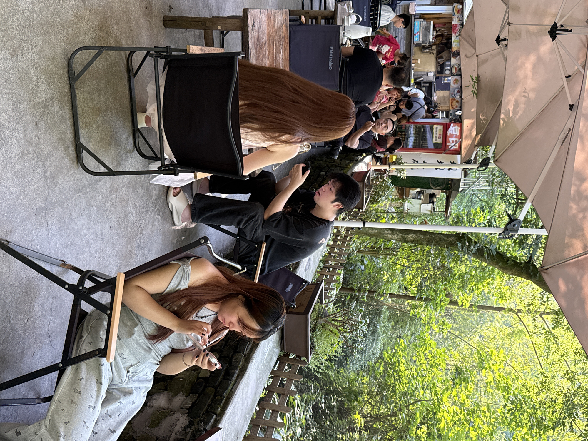 The image shows a casual outdoor scene, potentially a cafe or eatery in Beibei, China. Three people are visible, seated on portable chairs at separate tables. One woman wears a grey outfit and is looking at her phone. Another man is also looking at his phone. A third woman is sitting nearby. Other people are visible further in the background. There are tables and chairs set up for the customers. The environment includes greenery, indicating a natural or park-like setting with possible exposure to lush trees and plants. Parasols are used to cover customers from the sun.