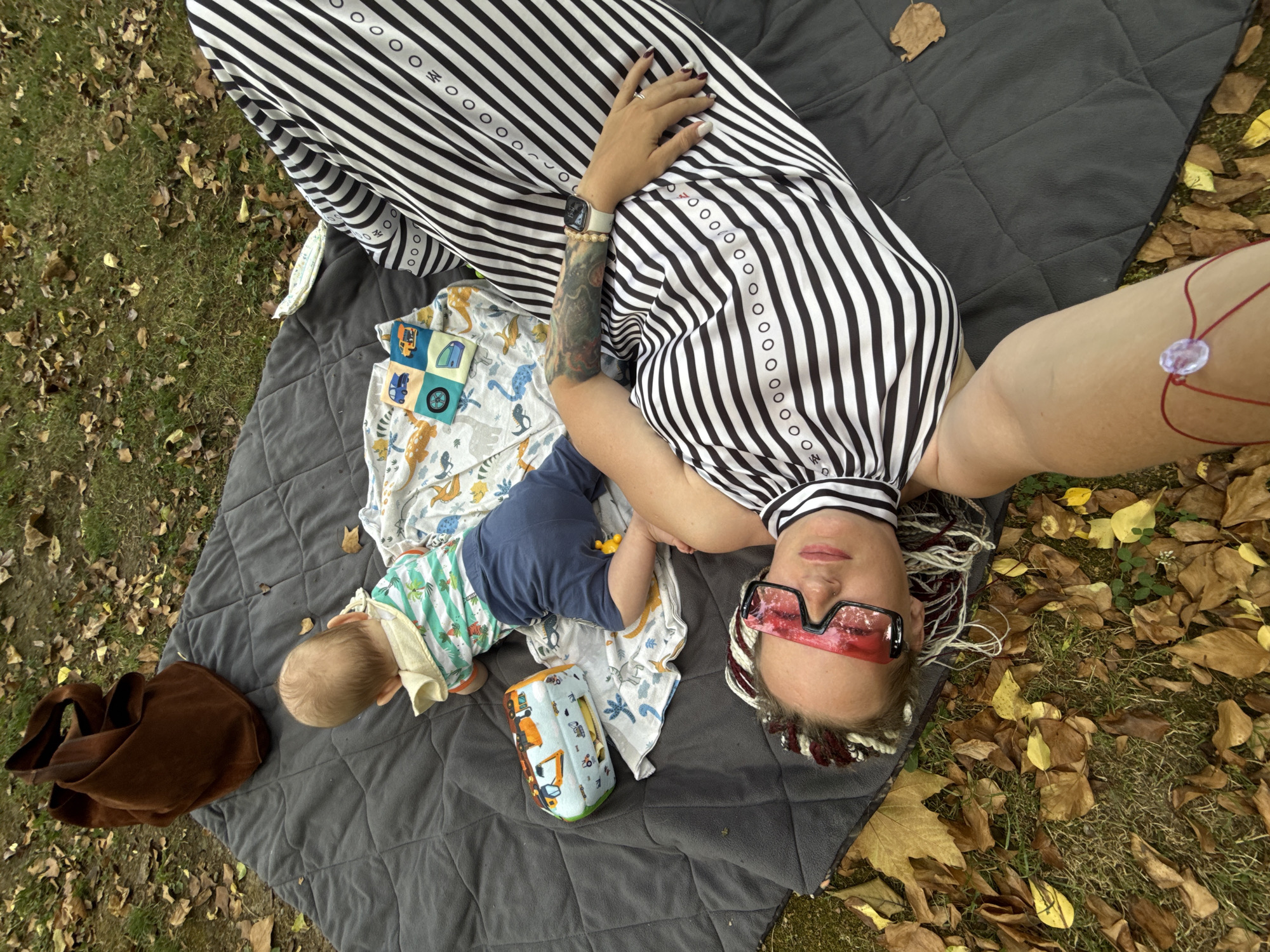 In Sochi, Russia, a woman with braided hair, wearing oversized red-rimmed sunglasses and a black and white striped dress, lies on a grey picnic blanket on the ground. Her tattooed arm rests on her stomach, and a watch is visible on her wrist. Beside her, a baby wearing a striped shirt and blue shorts is crawling. A brown leather bag, a soft toy with vehicle prints, and a blanket with dinosaur illustrations are also on the picnic blanket. Fallen autumn leaves are scattered on the grass surrounding the blanket. The image is taken from a high-angle perspective.