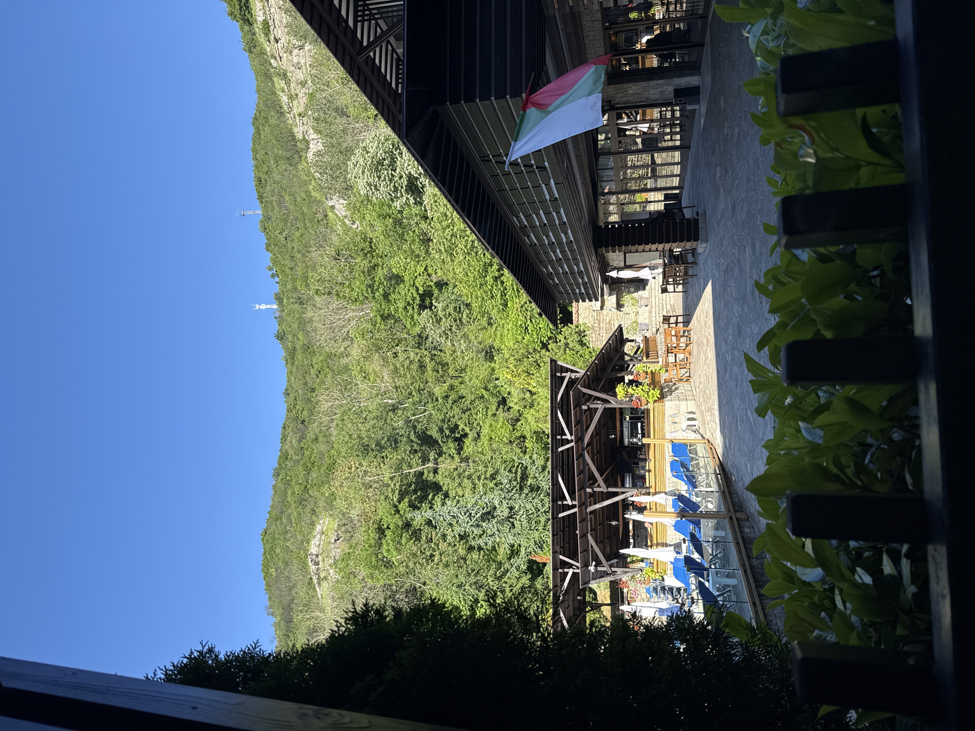 The image shows a view in Veliko Tarnovo, Bulgaria, encompassing a natural and architectural setting. A green, tree-covered mountain stretches upward under a clear blue sky. Two transmission towers are visible on the mountain. A wooden structure with a Bulgarian flag is near the top edge of the image. The lower half of the image features a partially shaded restaurant or terrace area with tables, chairs, and blue seating. Vegetation frames the view on the right side and lower edge. The image is oriented vertically.