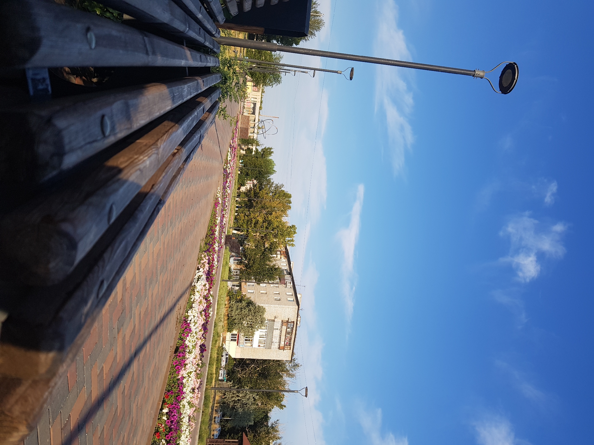 The image depicts a park-like setting with a paved walkway lined with flowerbeds and a wooden bench in the foreground. In the background, a multi-story residential building and trees are visible under a bright blue sky with scattered clouds. A street lamp stands to the left of the frame. The location is identified as Kurchatov, Kazakhstan. The scene appears to be an outdoor public space, possibly a promenade or a park, characterized by its clean and well-maintained appearance. There are no people or animals present in the frame. The overall impression is one of a serene and orderly urban environment.
