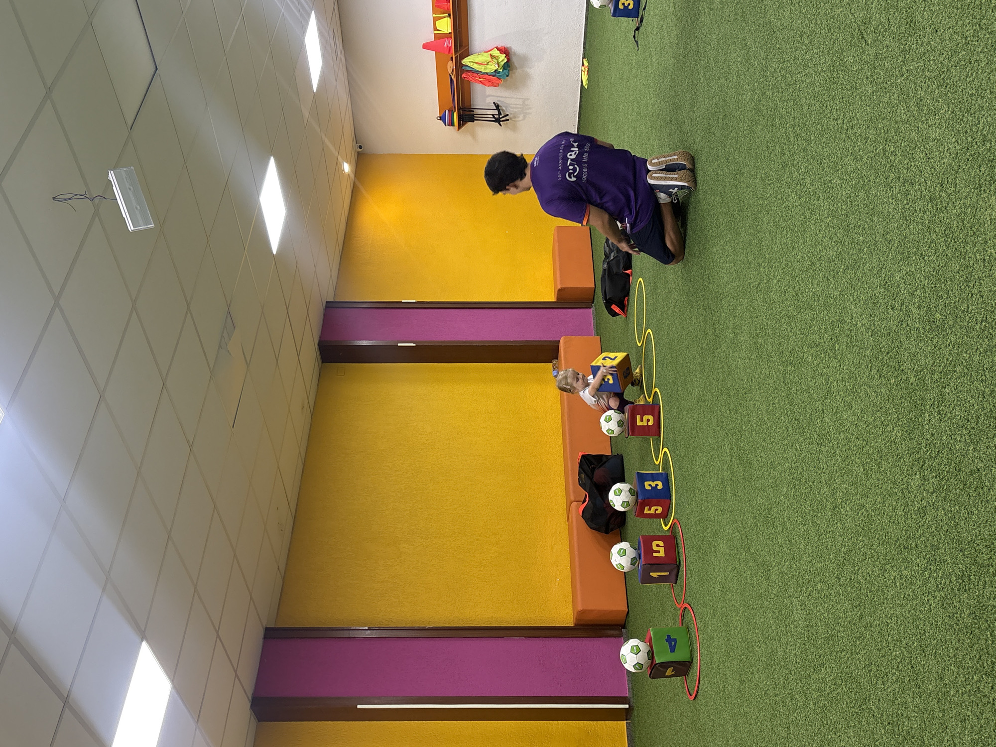 The image depicts an indoor children's sports facility, possibly a soccer training area, located in Valencia, Spain. The scene features a synthetic turf floor and vibrant yellow and orange walls. In the foreground, a young child is interacting with a large yellow cube. A male instructor, wearing a purple t-shirt, is kneeling on the turf, facing away from the camera and observing the child. Several soccer balls are placed on top of numbered blocks arranged on the floor, and yellow hoops are scattered around the activity area. A rack with various sports equipment, including cones and vests, is visible on a white wall in the background. The lighting appears to be artificial, with fluorescent panels on the ceiling.
