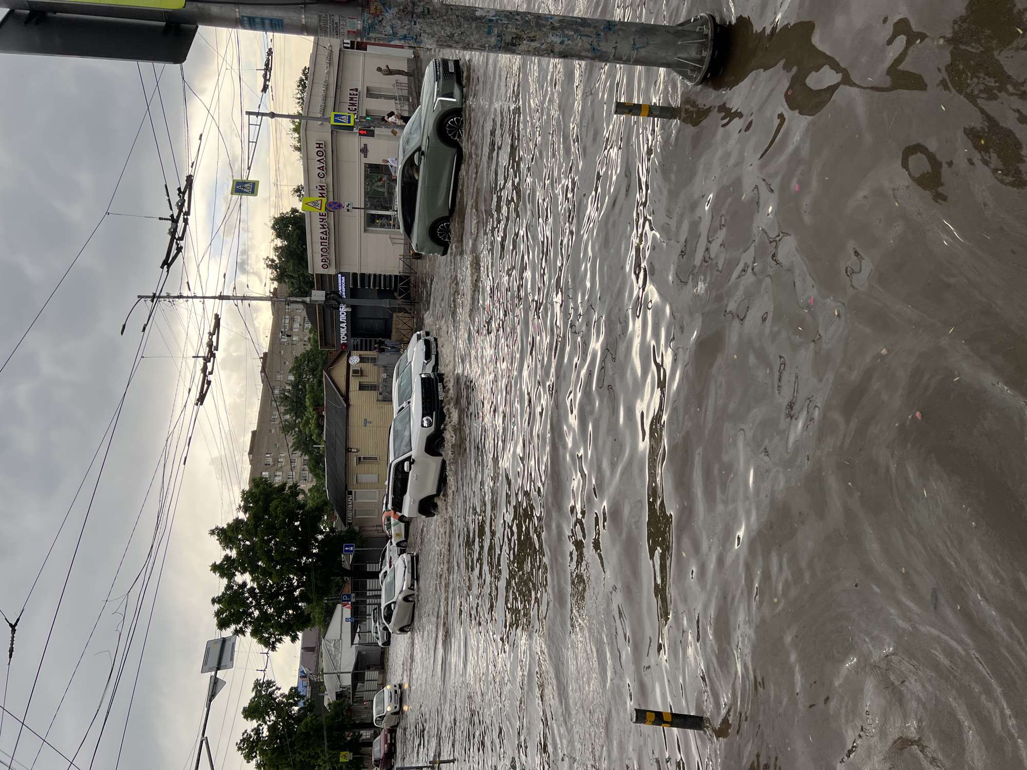 The image depicts a flooded street in Krasnodar, Russia, with several cars driving through the deep water. The sky is overcast with dark clouds, suggesting recent or ongoing rainfall. The street appears to be lined with buildings, some of which are commercial establishments with signage visible. The water level is high enough to submerge the lower parts of the vehicles, and ripples indicate the movement of the cars through the water. Pedestrians can be seen in the background, some walking near the flooded area. The scene suggests a disruption of normal city life due to the heavy flooding.