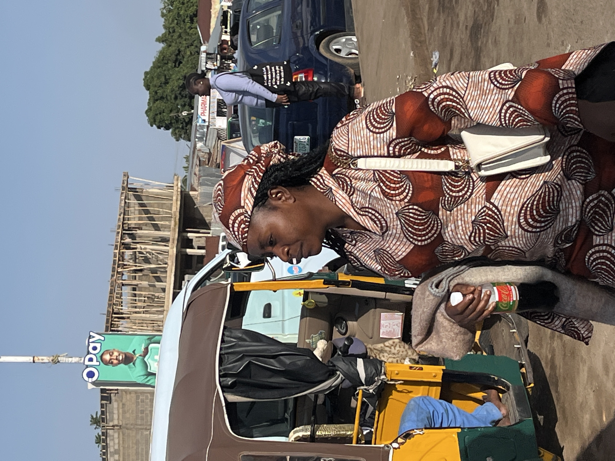 The image depicts a street scene in Gura Top, Nigeria, under a clear sky. In the foreground, a woman wearing a patterned headscarf and a matching long-sleeved dress walks towards the right of the frame. She is carrying a brown blanket and a bottle of Coca-Cola in her right hand, and a white handbag with a long strap is slung across her left shoulder. To her left, a yellow and brown auto-rickshaw is parked, with a person's legs clad in blue jeans visible inside. In the background, a man in a green shirt is featured on a large "OPay" advertisement mounted on a pole. Further back, construction is underway on a building with wooden scaffolding. A blue car is parked on the right, and a woman wearing a light blue shirt and dark pants stands beside it, holding a black tote bag. Various market stalls and other vehicles are scattered throughout the background, suggesting a bustling urban environment.