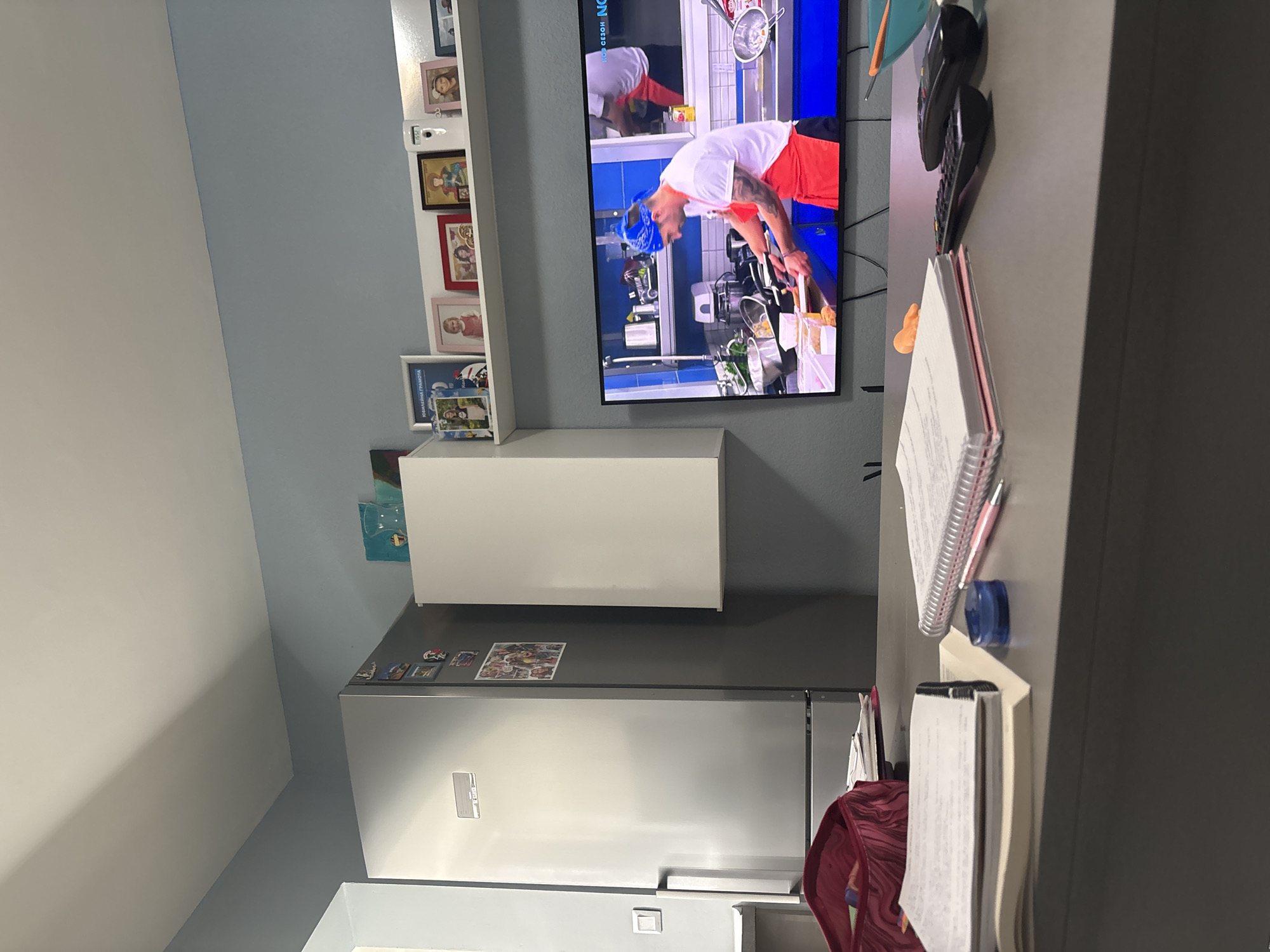 The image depicts an indoor scene, likely a kitchen or living area within a home in Sofia, Bulgaria. The room features walls painted in two tones: a light grey or white on the left and a light blue-grey on the right.
On the left side, a white floating shelf is mounted on the wall, displaying several framed photographs. These include portraits of adults and children, suggesting family photos. A small white device, possibly a security camera or smart home gadget, is also visible on the shelf. One framed item clearly shows the text "ОБРАЗЕЦ НА ГРАМОТА" (Model of Diploma/Certificate) in Cyrillic.
Dominating the center of the image is a large flat-screen television mounted on the wall. The TV is displaying what appears to be a cooking show. A male figure, likely a chef, is visible on screen, wearing a white shirt, a red apron, and a blue bandana on his head, actively preparing food in a modern kitchen setting. Text visible on the TV screen includes "НОВ СЕЗОН" (New Season) and "NOVO" (New), along with part of a channel logo. Below the television, a white rectangular wall-mounted cabinet or shelf is present.
To the right of the cabinet, a stainless steel refrigerator is visible, with a few small magnetic items or photos adhered to its surface. On the far right, part of a desk or counter is visible. Several notebooks or binders are stacked, along with a pink pen, a blue pen, and a blue magnetic clip. A red backpack or bag hangs from an unseen hook or chair just outside the frame. The overall impression is a functional and lived-in domestic space.