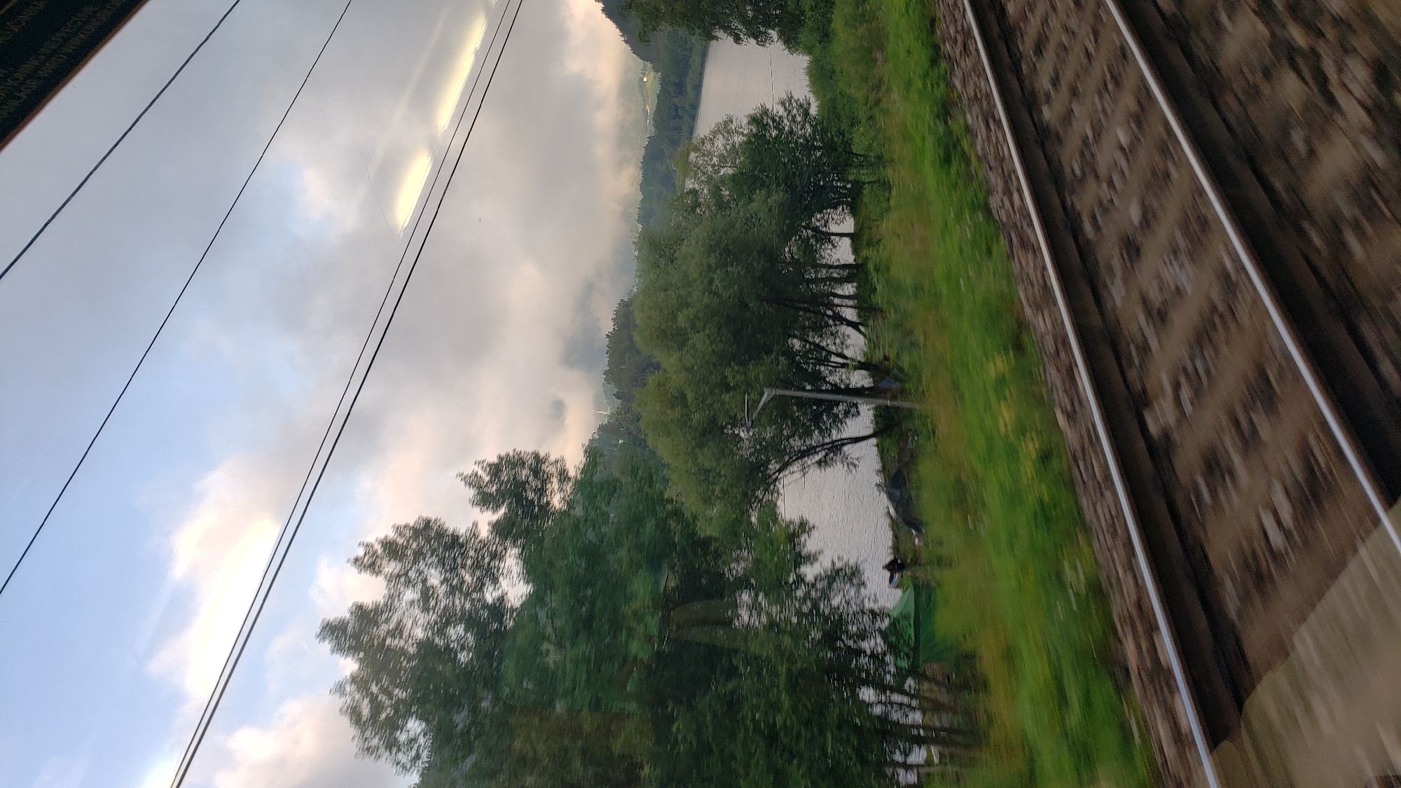 A view from a moving train shows railroad tracks in the foreground, blurred due to motion. Beyond the tracks, a body of water, likely a lake or river, is visible, bordered by a treeline and a grassy embankment. In the distance, hills and a cloudy sky are present. The scene appears to be in or around Margecany, Slovakia, a location known for its scenic landscapes and proximity to water bodies.