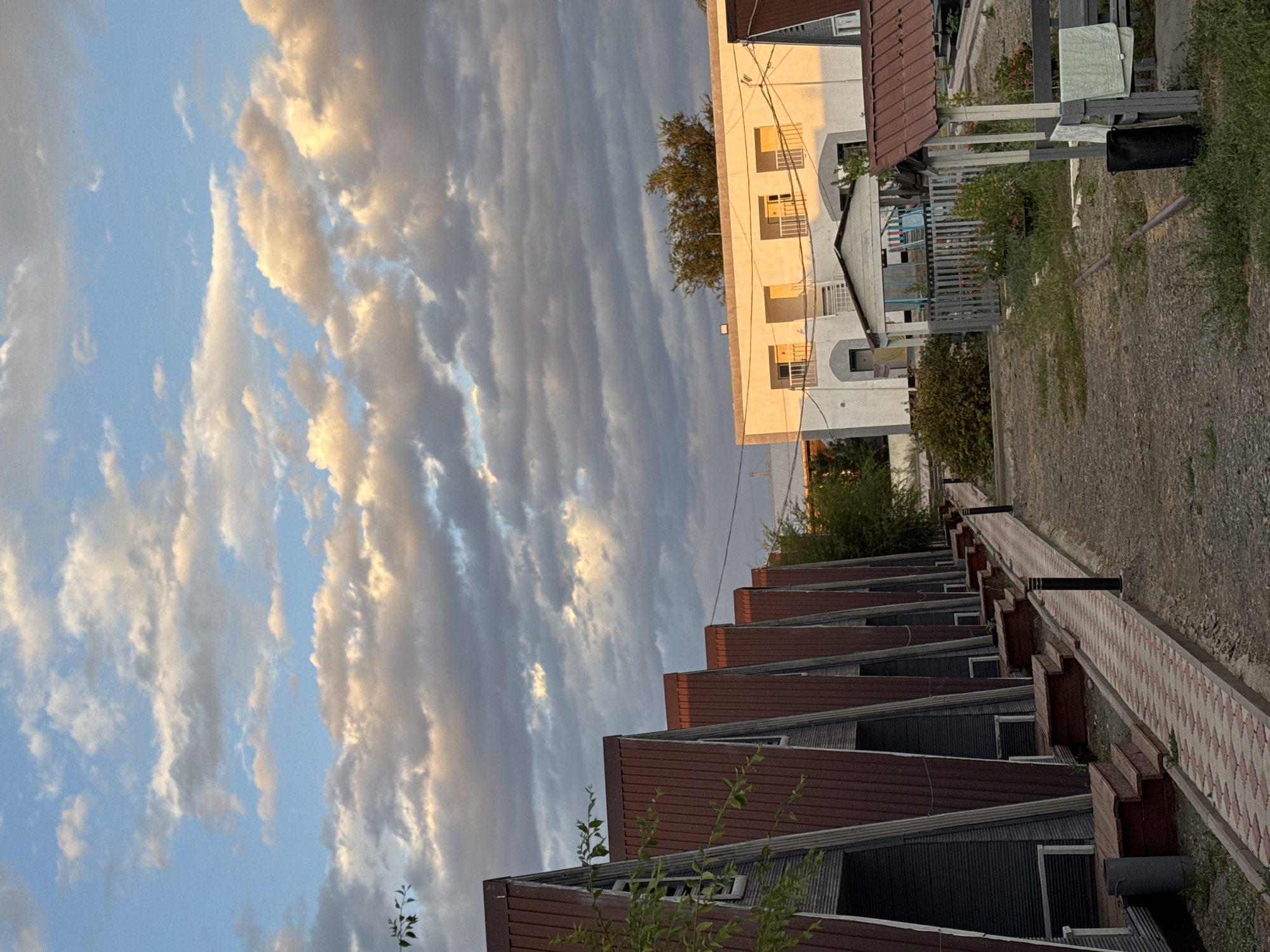 In Zharbulak, Kazakhstan, at sunset, a row of triangular, maroon-roofed structures with dark siding is visible from a dirt path. Each structure has a wooden deck and steps leading to its entrance. The path, paved with red and white interlocking bricks, is lined with lampposts. In the background, a white, two-story building with balconies and yellow-paned windows is illuminated by the setting sun. A small, open-air gazebo with a grey wooden fence and colorful flowers stands in front of the white building. The sky above is filled with dramatic clouds, tinged with orange and pink from the sunset. Vegetation, including trees and bushes, is scattered throughout the scene.