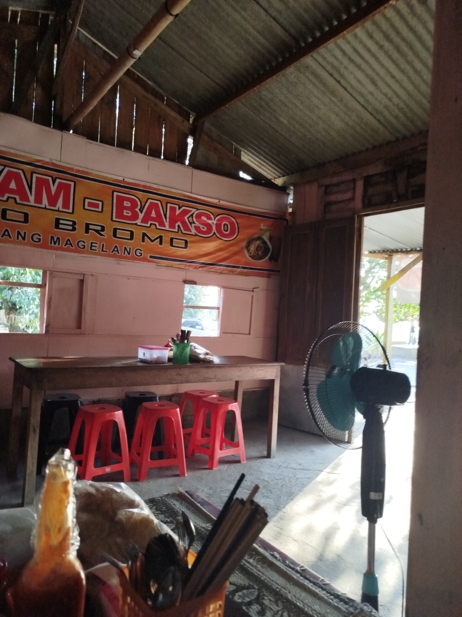 The image depicts the interior of a modest food stall or restaurant, likely in the city of Pojok, Indonesia. The setting appears rustic, with a corrugated metal roof supported by exposed wooden beams and vertical wooden planks. A large orange banner with red lettering is prominently displayed on the wall, reading "AM BAKSO" and below it, "O BROMO ANG MAGELANG". This suggests the establishment specializes in Bakso (meatball soup) and is possibly named "BaksoBromo" or related to the Bromo region and Magelang.
In the center of the room, a long wooden table is set up with several red plastic stools lined up in front of it. On the table, there's a white plastic container with a pink lid, a green cup holding chopsticks, and a small brown bag. Two windows are visible on the wall behind the table, allowing natural light to enter. One window looks out onto greenery, while the other offers a glimpse of a road with a parked vehicle.
To the right of the scene, a standing electric fan with a teal-colored motor and blades is positioned near an open doorway. The doorway leads to an outdoor area with more natural light and some foliage. In the foreground, partially out of focus, is a bottle with an orange-colored liquid, possibly sauce or chili, and a basket containing what appear to be chopsticks or skewers. The floor is concrete, with a patterned rug partially visible in the lower foreground. The overall impression is of a casual, local eatery.