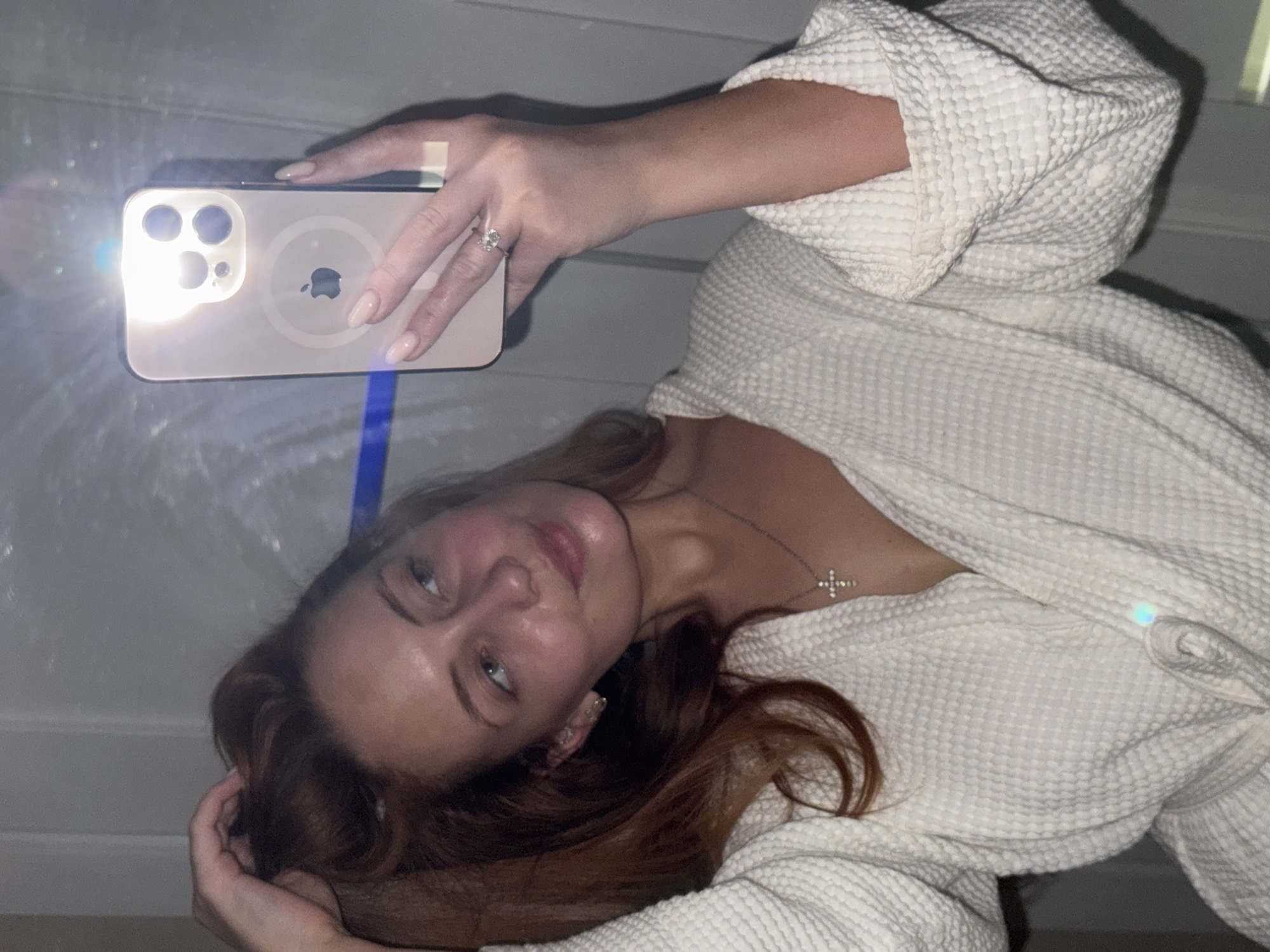 A woman with red hair is holding a gold iPhone and taking a selfie in an indoor setting. She is wearing a white waffle-textured robe and a delicate silver necklace with a cross pendant. She is wearing a ring on her finger. The phone's flash is illuminated. The background appears to be a simple white wall. The location is not discernible from the image.