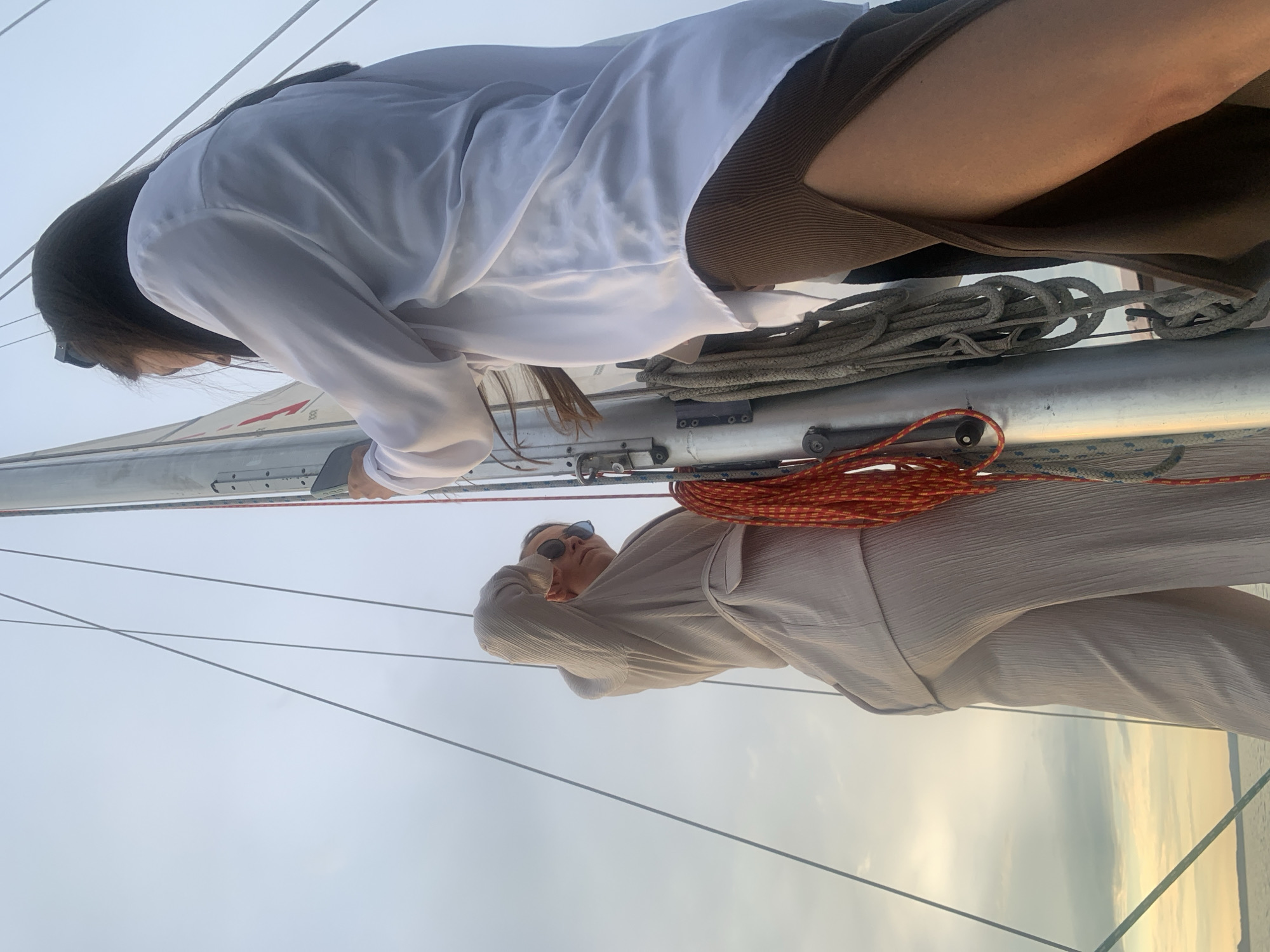 Two individuals are on a sailboat, likely in the city of Chaykovsky, Russia, given the context. The person on the left is wearing light-colored, textured pants and a matching top, with sunglasses perched on their head. They are looking towards the right, with their left arm raised. The person on the right is wearing a white, sheer button-down shirt and a brown, ribbed skirt with a high slit. They are holding a phone and appear to be taking a photo. The mast of the sailboat is central to the image, with various ropes attached. The background shows a cloudy sky with hints of sunset colors over a body of water, suggesting a coastal or riverine location consistent with Chaykovsky.
