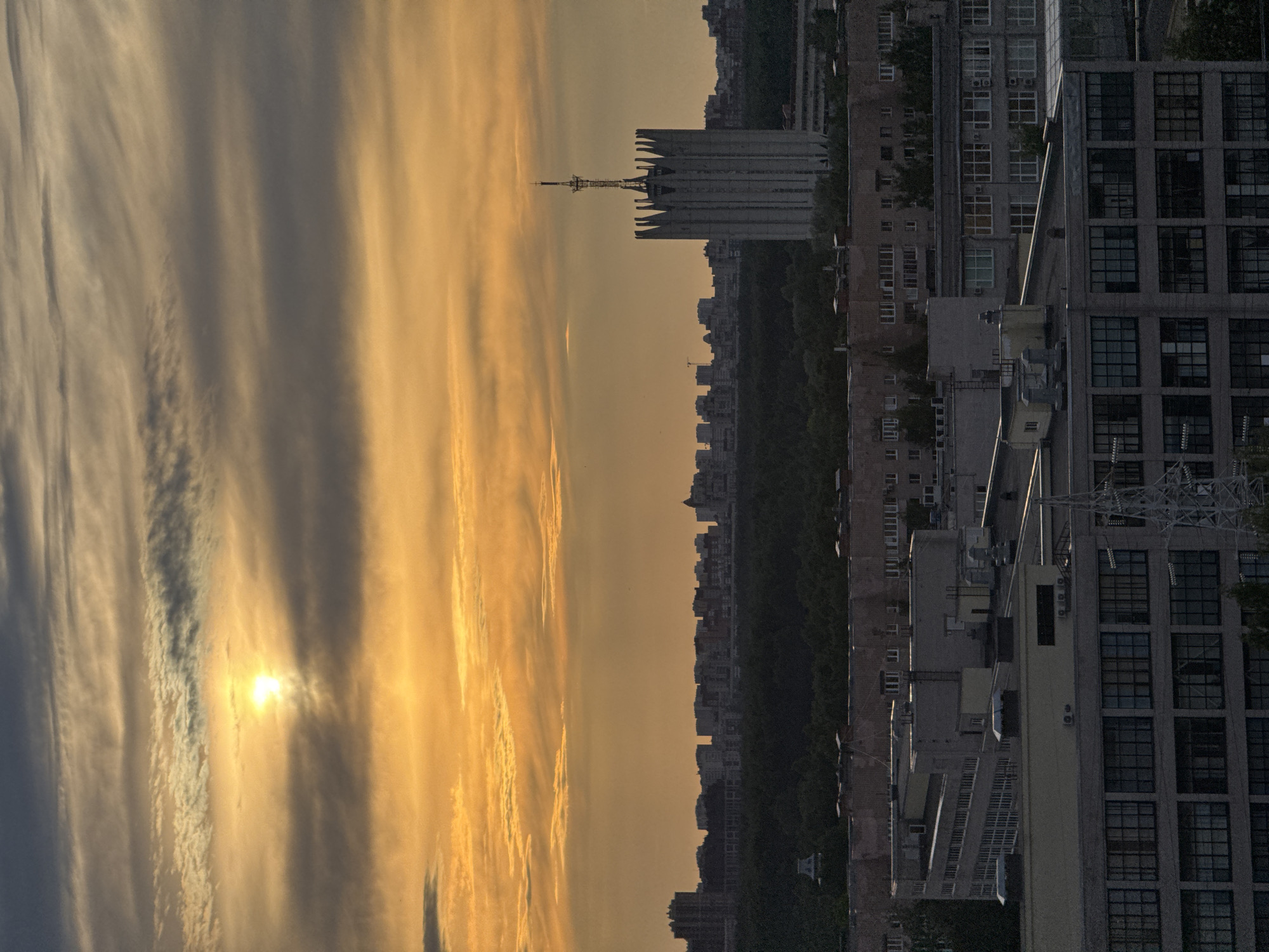 The image displays an urban skyline at either sunrise or sunset, viewed from an elevated position. The sky occupies the upper two-thirds of the frame, characterized by dramatic, streaky clouds. The sun, a brilliant, luminous disc, is visible on the left side, piercing through the clouds and casting an intense golden and orange glow across the sky. Patches of clouds remain in shadow, appearing in darker grey and bluish tones, creating a striking contrast with the sunlit areas.
Below the horizon, a sprawling cityscape is visible. A distinctive grey building with a unique, serrated or spiky roofline and a tall, slender antenna or spire stands out prominently in the mid-ground. This architectural feature is a key visual element against the colorful sky. Further in the distance, a dense line of darker buildings forms a silhouette, punctuated by a stretch of dark green trees.
In the foreground and mid-ground to the right, several multi-story buildings are more clearly discernible, featuring numerous windows, balconies, and air conditioning units. A large building on the far right has many dark, square windows, while a lighter-colored building sits adjacent to it. The prevailing light bathes many of these structures in shadow, highlighting their forms against the radiant sky. No people or specific activities are visible. The scene exudes a tranquil yet dramatic mood, typical of dawn or dusk over a major city, fitting the provided context of Russia. No visible text is present in the image.