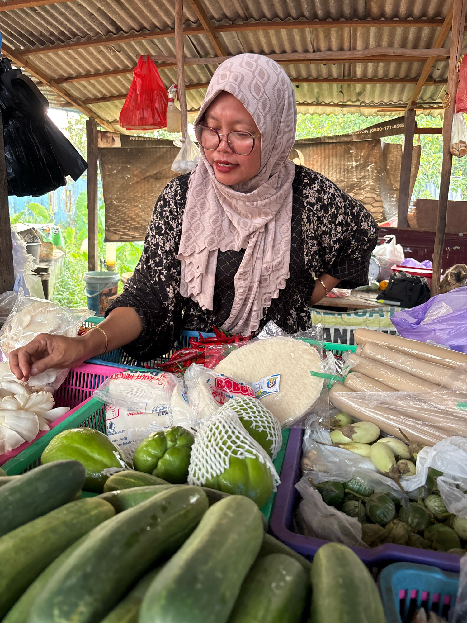 A middle-aged Indonesian woman wearing a light pink hijab and glasses is seen at a market stall in Banjarmasin, Indonesia. She is looking down and appears to be interacting with the items in her stall. The stall is covered by a corrugated metal roof supported by wooden beams. Various fresh produce, including cucumbers, green chilis, and what appears to be mushrooms, are displayed in plastic baskets. Packaged goods are also visible. In the background, there are other market elements such as bags, buckets, and foliage, suggesting an outdoor or semi-outdoor market setting. A sign with text, partially obscured, is visible behind her.