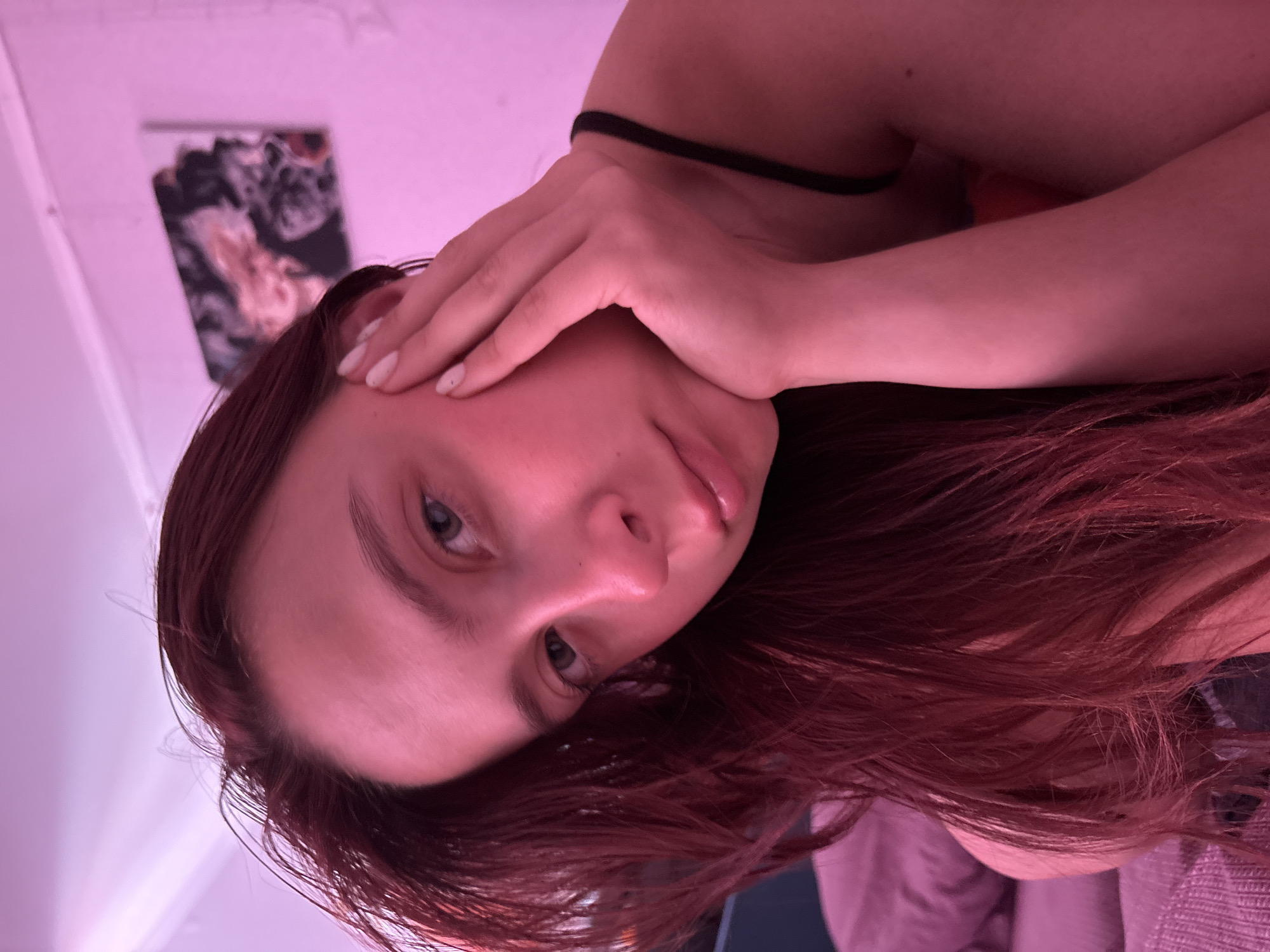A young woman with long, dark red hair is shown in a close-up selfie. She is indoors, illuminated by pink lighting, possibly from a lamp or artificial light source. Her right hand is resting on her cheek, and her left hand is near her face. She is wearing a black strap, likely from a tank top or bra. In the background, a framed piece of abstract art with swirling colors is visible on a white wall, and a pillow or bedding can be seen to her left. The overall impression is one of a casual, intimate moment captured at home. The location of Прикамский, Russia, cannot be determined from the image alone.