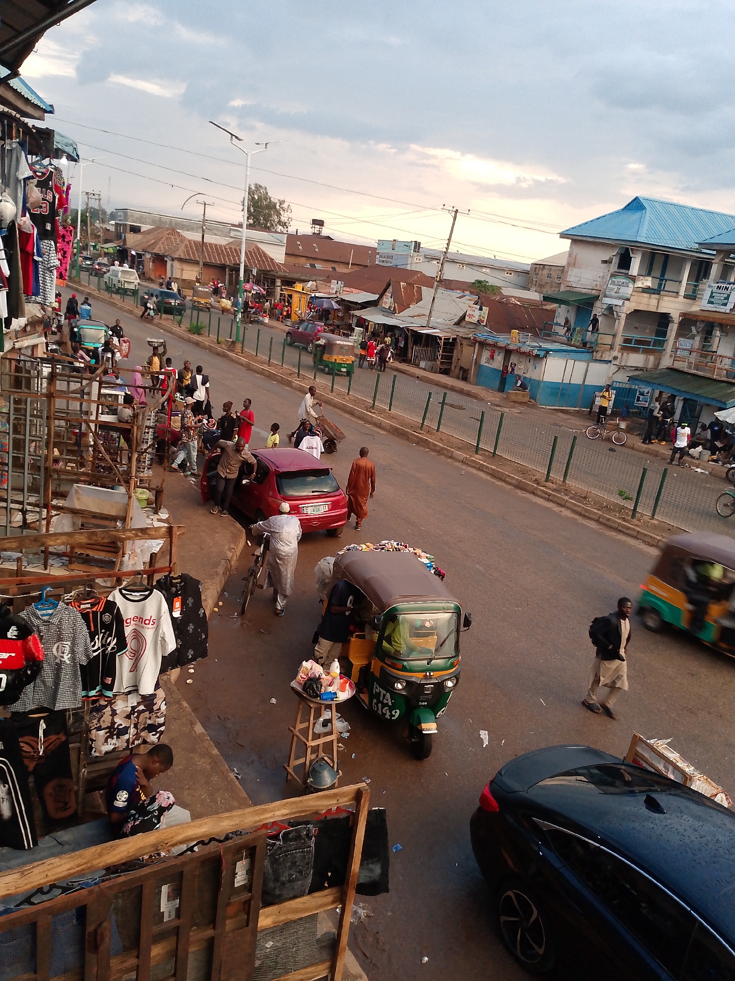 This image captures a bustling street scene in Bukuru, Nigeria, likely in the late afternoon or early evening under an overcast sky. The elevated viewpoint looks down upon a lively street bordered by market stalls and various buildings.
In the foreground, an open-air market stall displays an assortment of clothes, including t-shirts with "legends," "9," and a basketball jersey featuring "BULLS 23." A person is seen sitting, inspecting merchandise. A partial view of a dark-colored car is visible in the bottom right.
The street below is active with pedestrians and vehicles. A red car is parked with people gathered around it, one person entering or exiting. A green and yellow motorized tricycle, commonly known as a "keke napep," is clearly visible with the license plate "PTA 6149," and another, blurrier tricycle is further down the road. People in various attire walk along the pavement and in the street, including a man in a brown traditional robe and another in a light-colored outfit with a dark jacket.
The background features a mix of buildings, some with corrugated iron roofs indicative of local architecture, and a more prominent building with a striking blue roof. A green barrier fence separates the pedestrian area from the roadway. Power lines and a modern streetlight are visible against the cloudy sky. A sign reading "Home of NIN" can be seen on a building in the background. The scene conveys an atmosphere of everyday commerce and busy urban life.