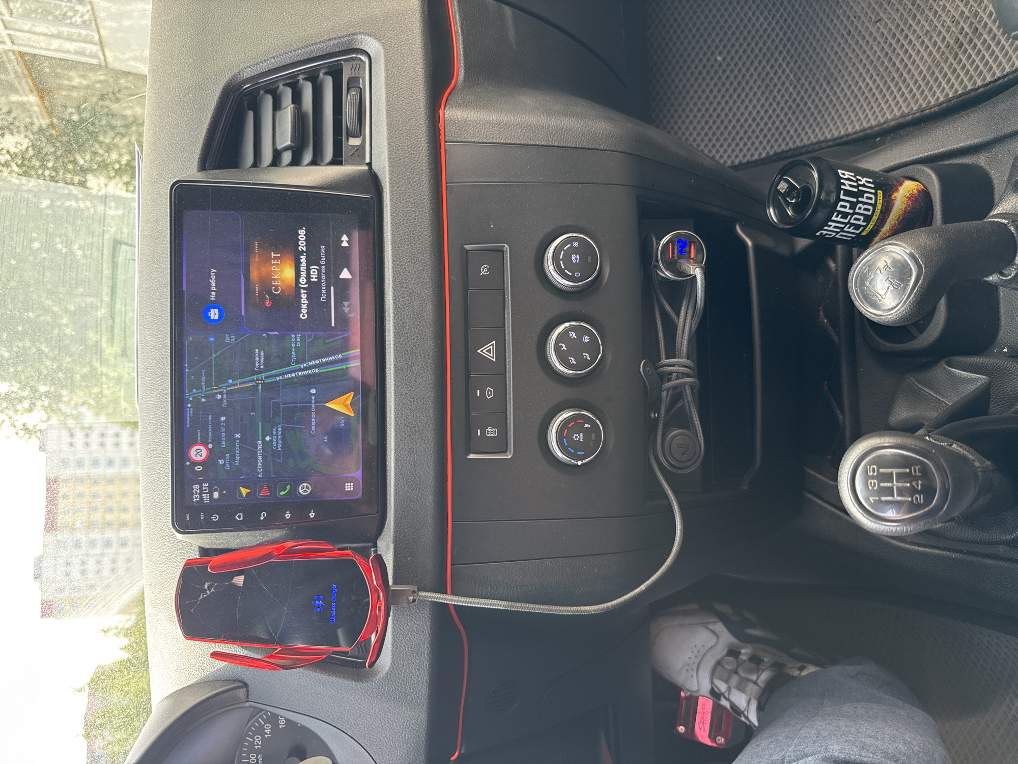 The image is a close-up view of the interior of a car, specifically the dashboard and center console. A modern infotainment screen is prominently displayed, showing a navigation map of a city, with a red indicator marking a location labeled "На работу" (To Work). Below the navigation, music playback is indicated, with "Секрет (Фильм, 2006, HD)" playing. A red wireless phone charger is mounted to the left of the screen, holding a black smartphone. The center console features climate control knobs, hazard lights, and a cigarette lighter adapter with a USB port connected to charging cables. A manual gear shifter is visible, with gears labeled "1, 3, 5, R, 2, 4". A can of "Энергия первых" (Energy of the First) energy drink rests in a cup holder next to the gear shifter. The floor mat is a textured gray. The exterior view through the windshield shows a blurred image of apartment buildings.
Based on the context provided (the Russian text on the screen and the energy drink can), this scene is likely located in Russia. The specific mention of Usinsk, Russia, is not directly visible in the image itself, but it can be inferred as a potential location for this photograph. The image captures a typical in-car environment, suggesting the vehicle is either parked or in motion.
