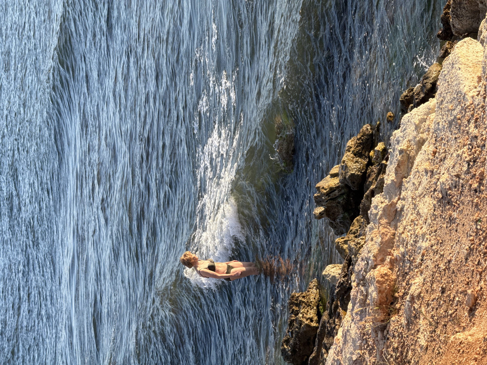 A woman in a black bikini stands in the shallow ocean water, facing away from the camera and towards a breaking wave. She has short blonde hair. The water is a deep blue with white foam from the wave. The foreground is a rocky, sandy shoreline. The overall lighting suggests it is either sunrise or sunset.