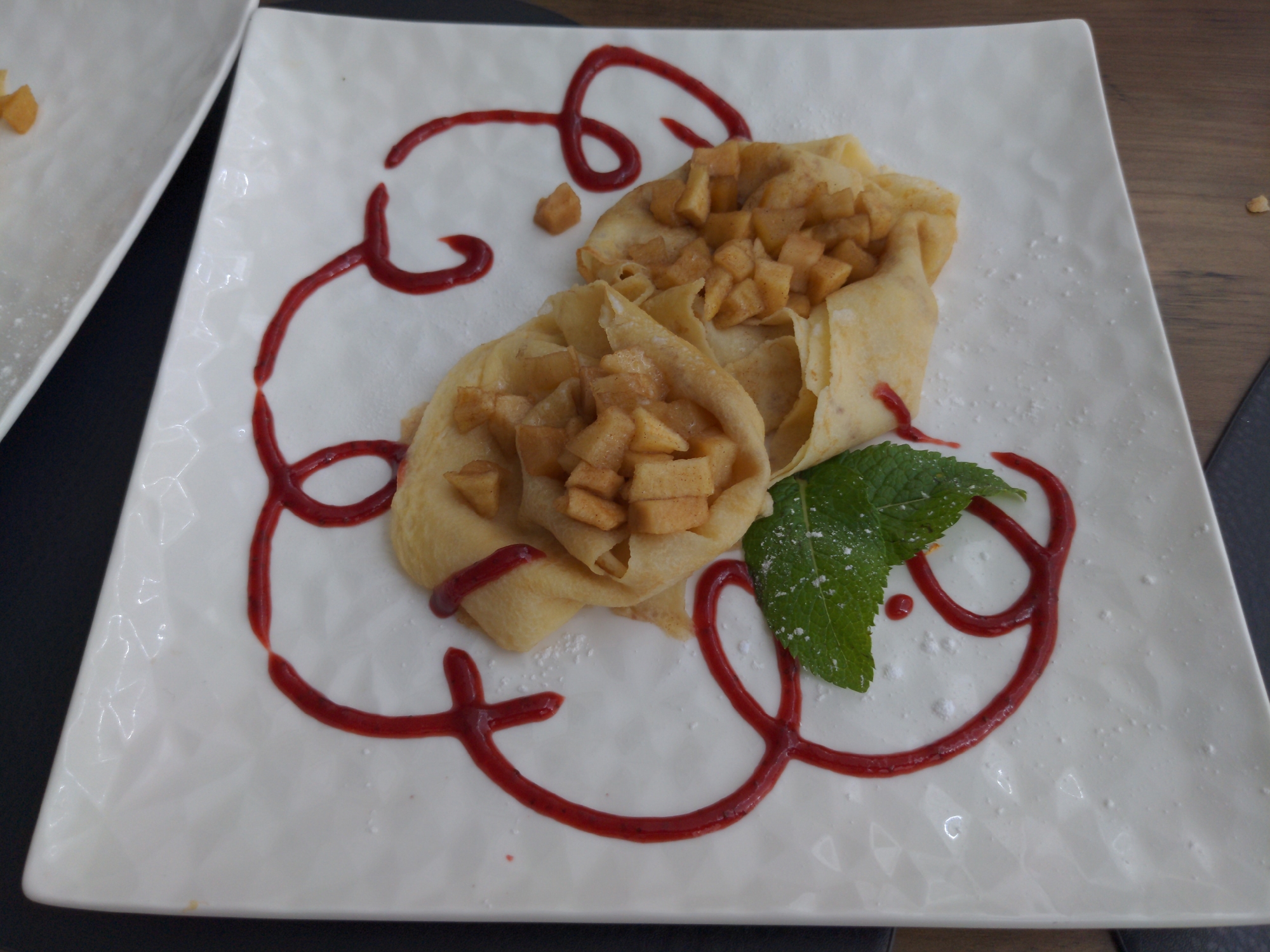 A close-up shot shows a dessert on a white, patterned plate. The dessert consists of two crepes filled with diced apples. The crepes are drizzled with a red sauce that forms a swirl pattern on the plate. A sprig of mint is placed on top of the crepes, and a light dusting of powdered sugar is visible on the plate and the mint. The scene is set on a wooden table. This image does not provide any information about the city of Holubyne, Ukraine.