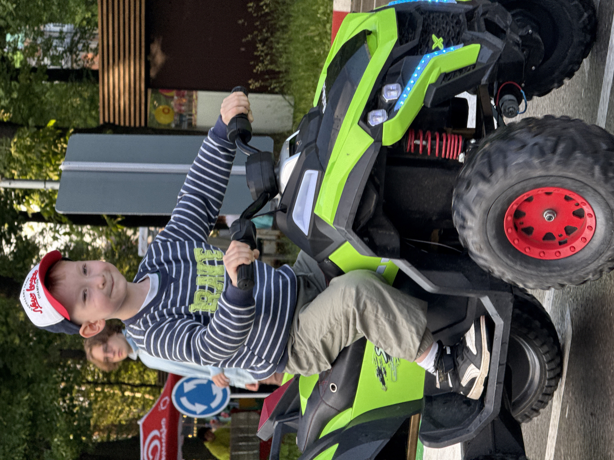 A young boy wearing a striped long-sleeved shirt and a red and white baseball cap is riding a green and black ATV. He is looking at the camera and smiling. The ATV has large, rugged tires with red rims. The boy is wearing white socks and black and white sneakers. Behind the boy, a person with blonde hair wearing glasses and a light blue shirt is visible, as well as a red tent and a circular traffic sign with blue arrows. The scene appears to be outdoors in a park or recreational area, likely in the city of Perm, Russia, given the context. The background shows trees, a lamppost, and a building with wooden siding.