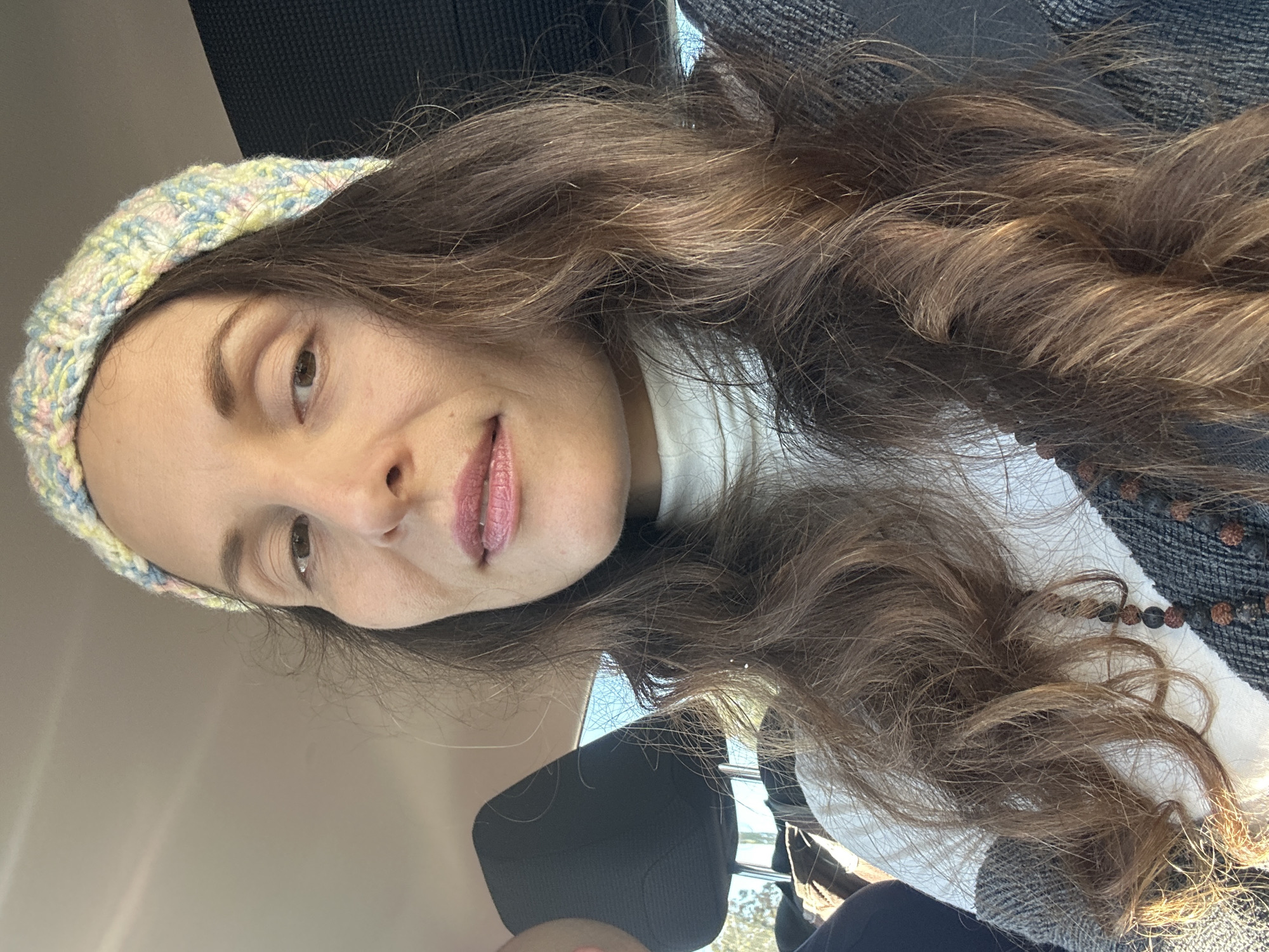 A woman wearing a pastel-colored knitted headband and a white turtleneck is captured in a close-up, selfie-style shot inside a vehicle. Her long, brown, wavy hair cascades over her shoulders. She is wearing subtle makeup, with pink lipstick on her lips. A beaded necklace, with dark, round beads, is visible around her neck. The background shows parts of the car's interior, including a headrest and a textured seat. Sunlight streams in from the right side of the frame, illuminating her face and hair. The overall impression is of a casual, candid moment.