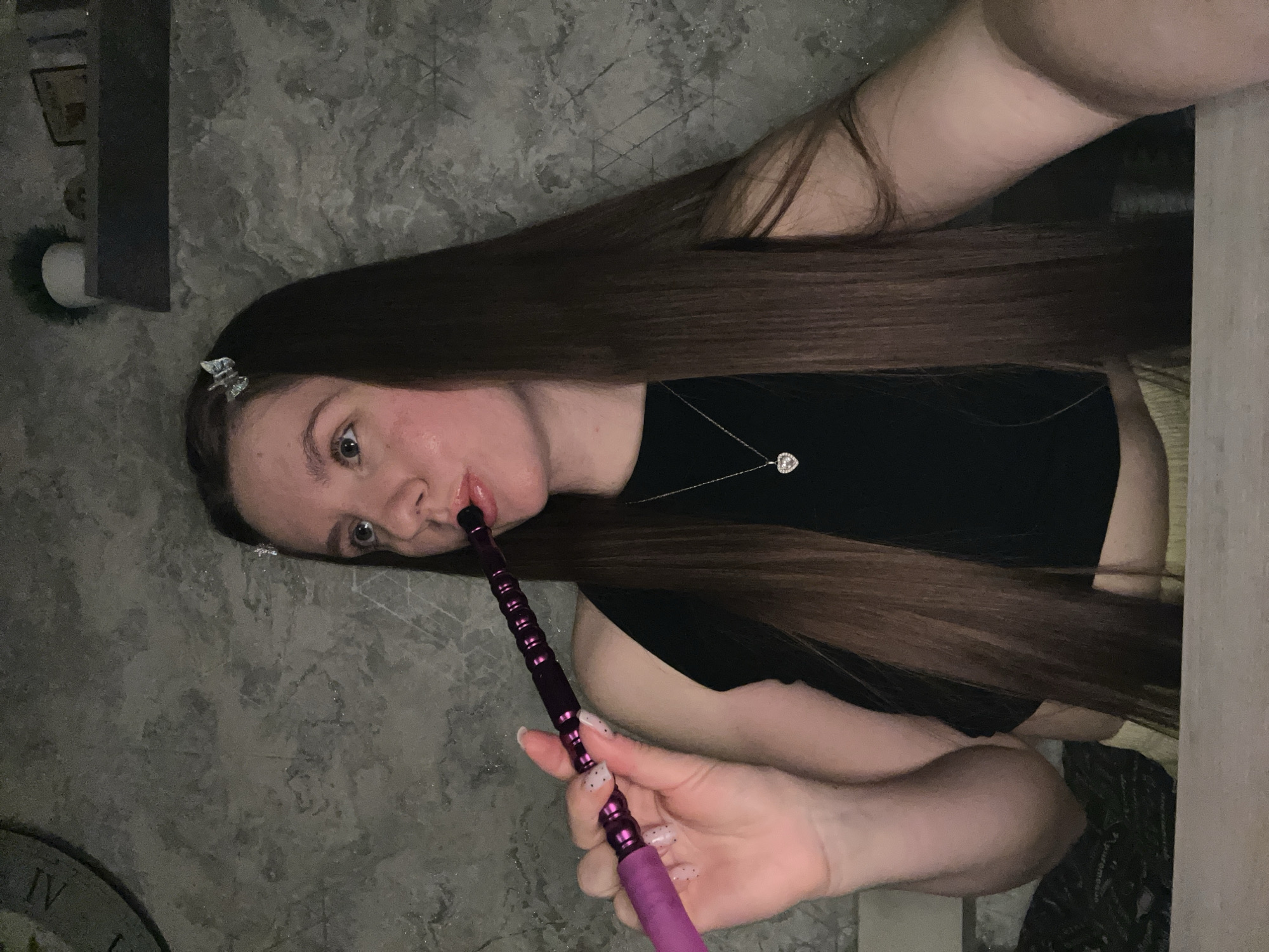 A woman with long, brown hair is holding and smoking a hookah. She is wearing a black top and a necklace. The background consists of a wall with a grey, marbled pattern. A portion of what seems to be a clock face can be seen in the lower-left corner. There is no definitive evidence that the location is Klin, Russia.