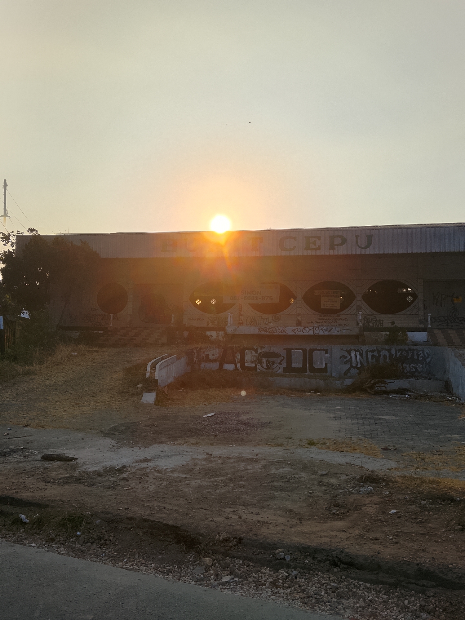 A medium shot captures a sunlit building in Ngelo, Indonesia, during what appears to be a sunset or sunrise. The sun, a bright orb, is positioned centrally behind the structure, casting a warm glow and lens flare across the scene. The building, with a weathered facade, displays the words "BU CEP" in large, dark lettering across its upper section. The front of the building features a row of oval-shaped openings, some of which are boarded up or obscured. The area in front of the building is unkempt, with dry grass, dirt, and debris scattered across the ground. Graffiti adorns the lower parts of the building and a concrete wall in the foreground, adding a layer of urban decay to the environment. The overall impression is one of an abandoned or disused commercial space bathed in the soft, diffused light of the sun.