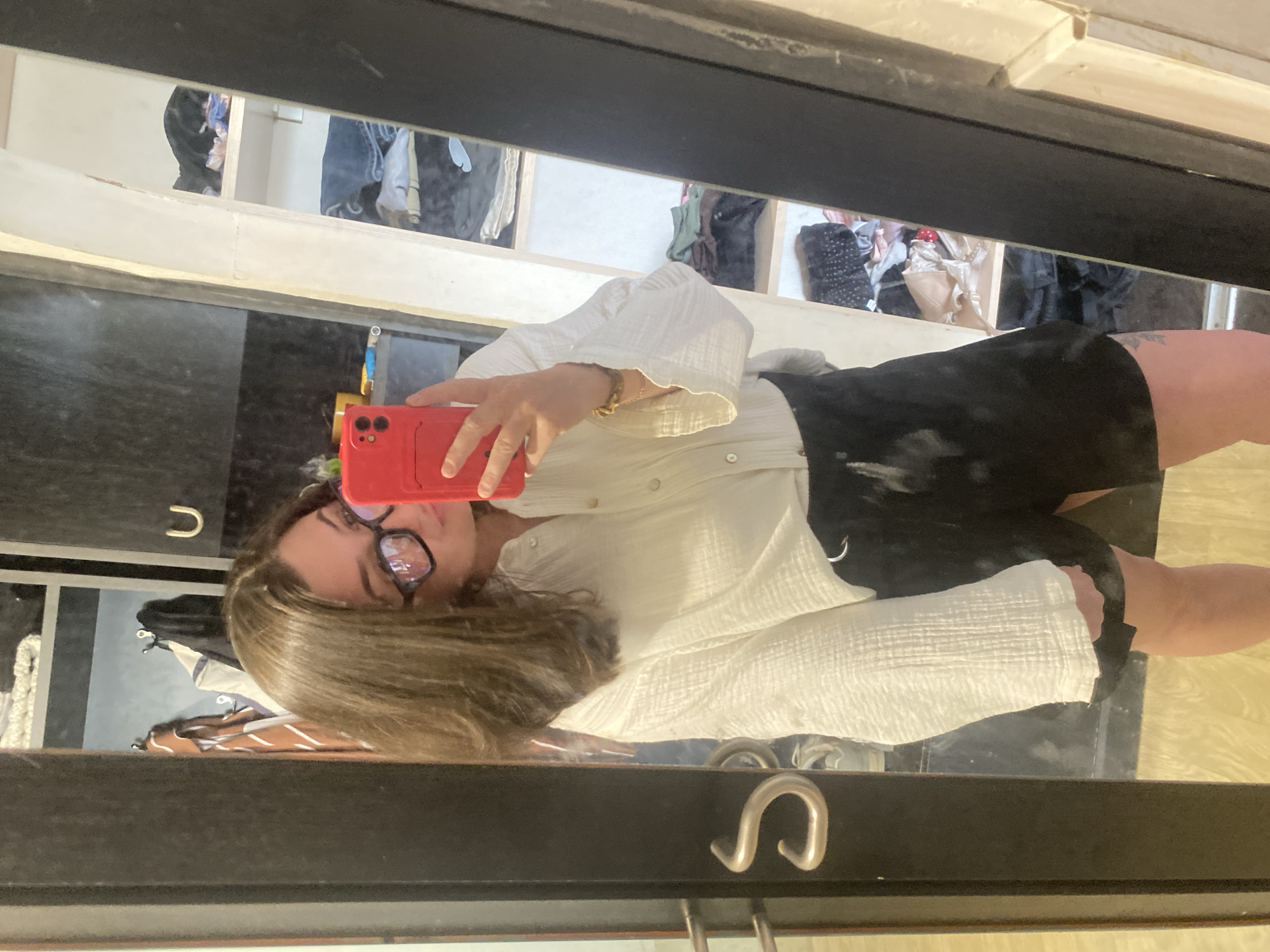 A person is taking a selfie in a mirror. The person has shoulder-length brown hair and is wearing glasses, a white shirt, and black shorts. A red phone is visible in the person's hand. The reflection in the mirror shows a closet with clothes hanging inside. The location is not provided in the image.