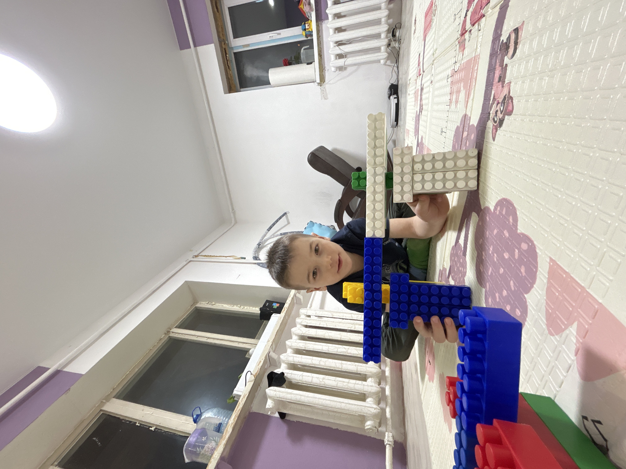 A young boy is lying on a play mat in a room in Rusalevka, Russia. He is holding a structure made of large, colorful plastic building blocks. The blocks are predominantly blue, red, yellow, green, and white. The room has a window, a white radiator, and a light-colored ceiling.