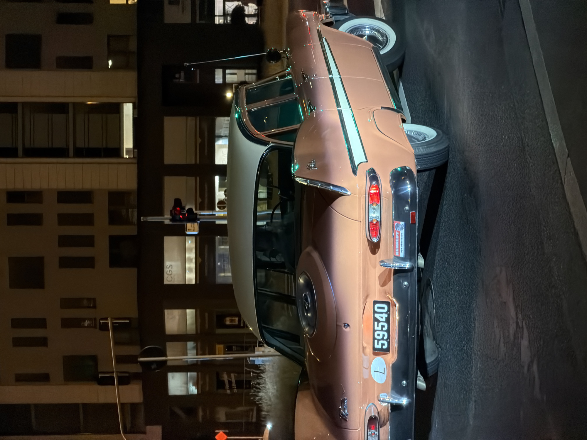 A salmon-pink colored vintage car, possibly a 1959 Plymouth Fury, is parked at night on a wet asphalt road. The car has a two-tone cream and salmon-pink roof, a chrome bumper, and a license plate that reads "L 59540". On the right side of the bumper, there is a "Kennedy for President" sticker. In the background, a building with illuminated windows and architectural details is visible. The streetlights cast a warm glow on the scene. The location is Esch-sur-Alzette, Luxembourg.