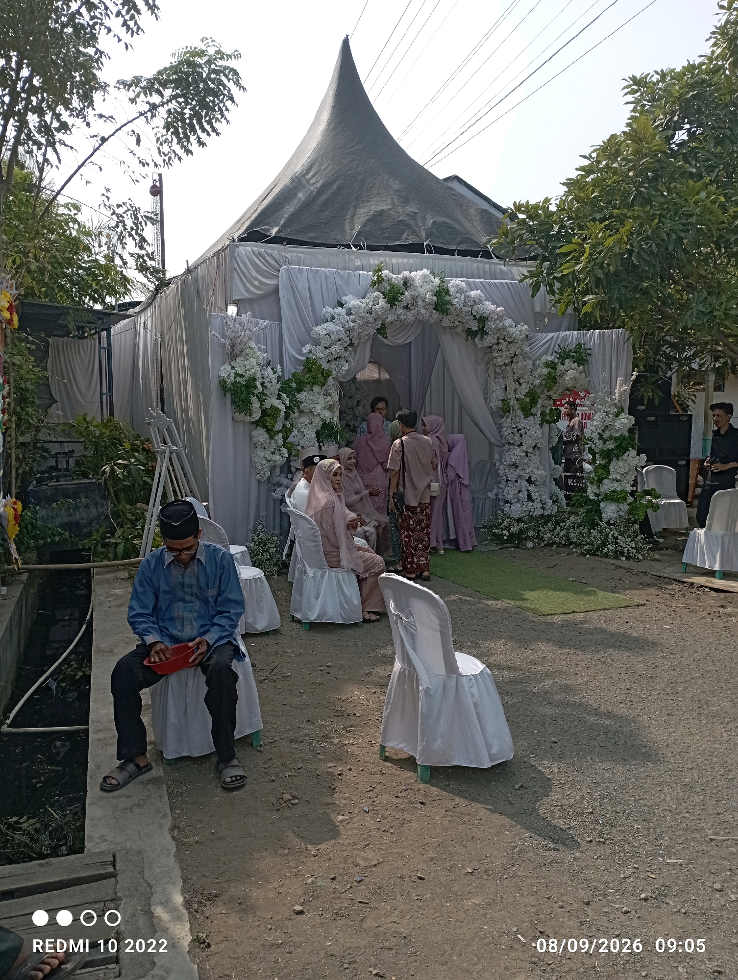 This image depicts an outdoor event, likely a wedding reception, set up in Dukuhseti, Indonesia. The scene features a large, peaked tent with white fabric draping, adorned with extensive floral arrangements, primarily white flowers. In the foreground, a man wearing a blue shirt, black pants, and a black cap is seated on a white-covered chair, holding a red bowl. Several other white-covered chairs are visible throughout the scene. Behind the main tent structure, other individuals are seen mingling, dressed in traditional attire, with women in pastel-colored hijabs and dresses. A green carpet or mat is laid out in front of the floral archway. The ground is a mixture of dirt and gravel. Trees and foliage frame the event space. The overall atmosphere suggests a warm, sunny day.