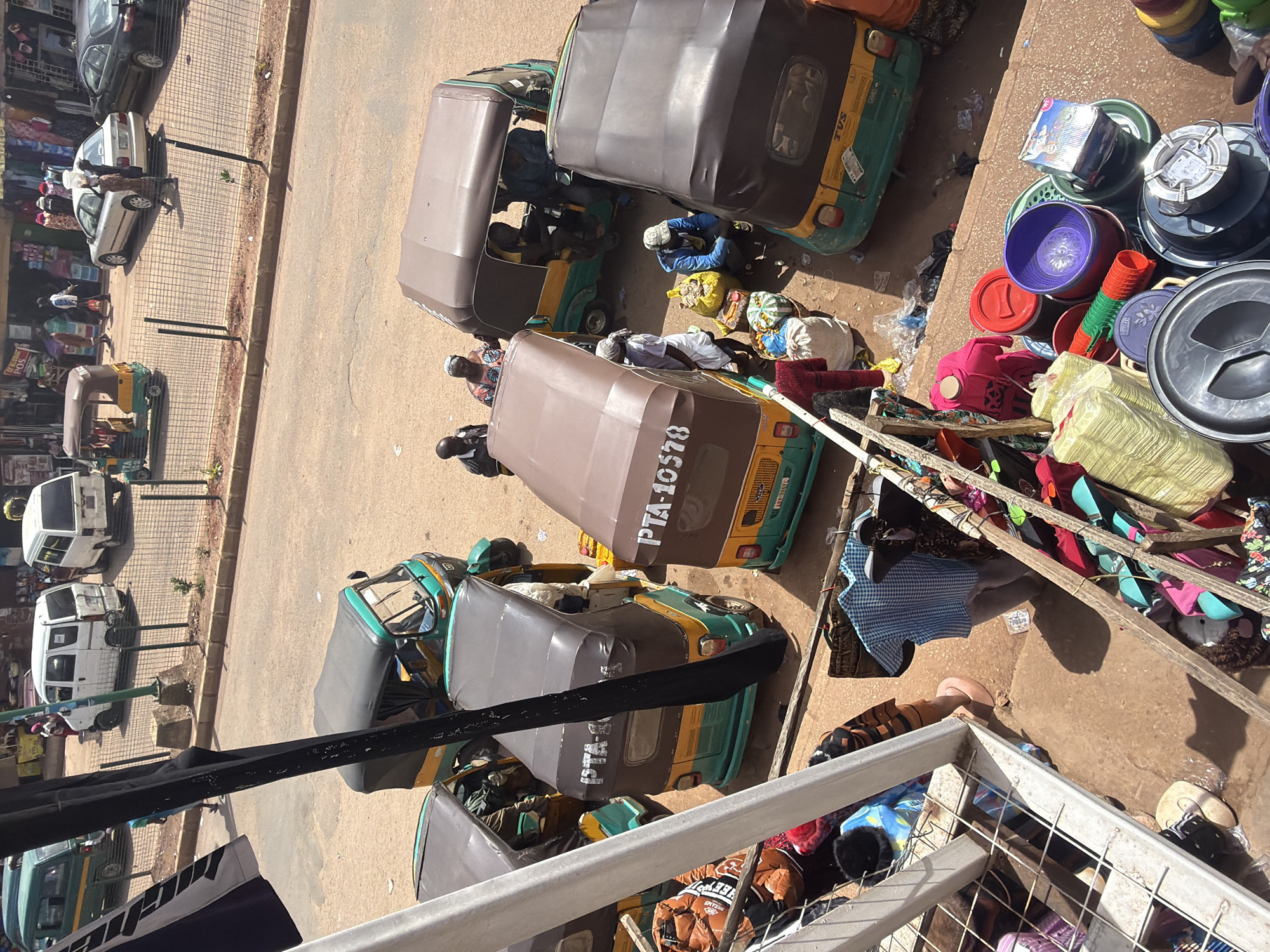 The image depicts a bustling street scene in Bukuru, Nigeria, likely during daylight hours, suggested by the strong shadows. Several auto-rickshaws, also known as *keke napeps*, are parked along a dirt road adjacent to a paved walkway. Some of these rickshaws are occupied by people, while others appear to be waiting for passengers or goods.
On the walkway, there are various market stalls or informal vendors displaying a range of items. These include stacks of colorful plastic bowls and containers, bundles of cloth, and what appear to be bags of goods. The overall impression is one of a busy commercial area where people are engaged in trade and transportation.
Notable details include the presence of multiple rickshaws, some with their roofs up, others with them down. The ground is a mix of dirt and what looks like a paved area. The objects for sale are varied, with plasticware and textiles being prominent. There is also some visible litter, indicating a lively but perhaps not meticulously clean environment.
Text is visible on some of the rickshaws, including "PTA-10578" and "TVS". This suggests registration numbers for the vehicles and potentially the manufacturer. The scene conveys a sense of everyday life and commerce in Nigeria, with a focus on local transportation and market activities. The atmosphere appears active and engaged.