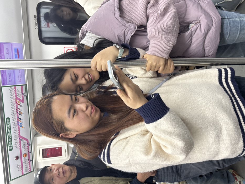 An indoor scene depicts passengers inside a modern subway train car on Chongqing Rail Transit Line 6, operating in or around Longxi Subdistrict (龙溪街道), China. The primary focus is on two East Asian females standing in the foreground. The female on the left, with long brown hair and wearing a cream-colored sweater with navy blue striped cuffs and hem, holds a light-colored smartphone horizontally in both hands, looking down at the screen with a slight smile. A light blue shoulder strap of a bag is visible across her body. Adjacent to her on the right, a second East Asian female with dark hair smiles, looking towards the smartphone screen. She is dressed in a lavender puffer vest over a lighter purple long-sleeved top and light blue jeans, with her right hand gripping a vertical polished metal pole. A light blue wristwatch is visible on her left wrist. To the far left, an older East Asian male in a dark cap and jacket is partially visible, looking forward. The subway car interior is clean and well-lit, with white walls. Above the passengers, a detailed route map for "重庆轨道交通6号线线" (Chongqing Rail Transit Line 6) is displayed, alongside a digital information screen showing Chinese text and the word "Approaching." The train car number "0602025B" is visible above a closed door in the background, where reflections of other passengers are discernible.