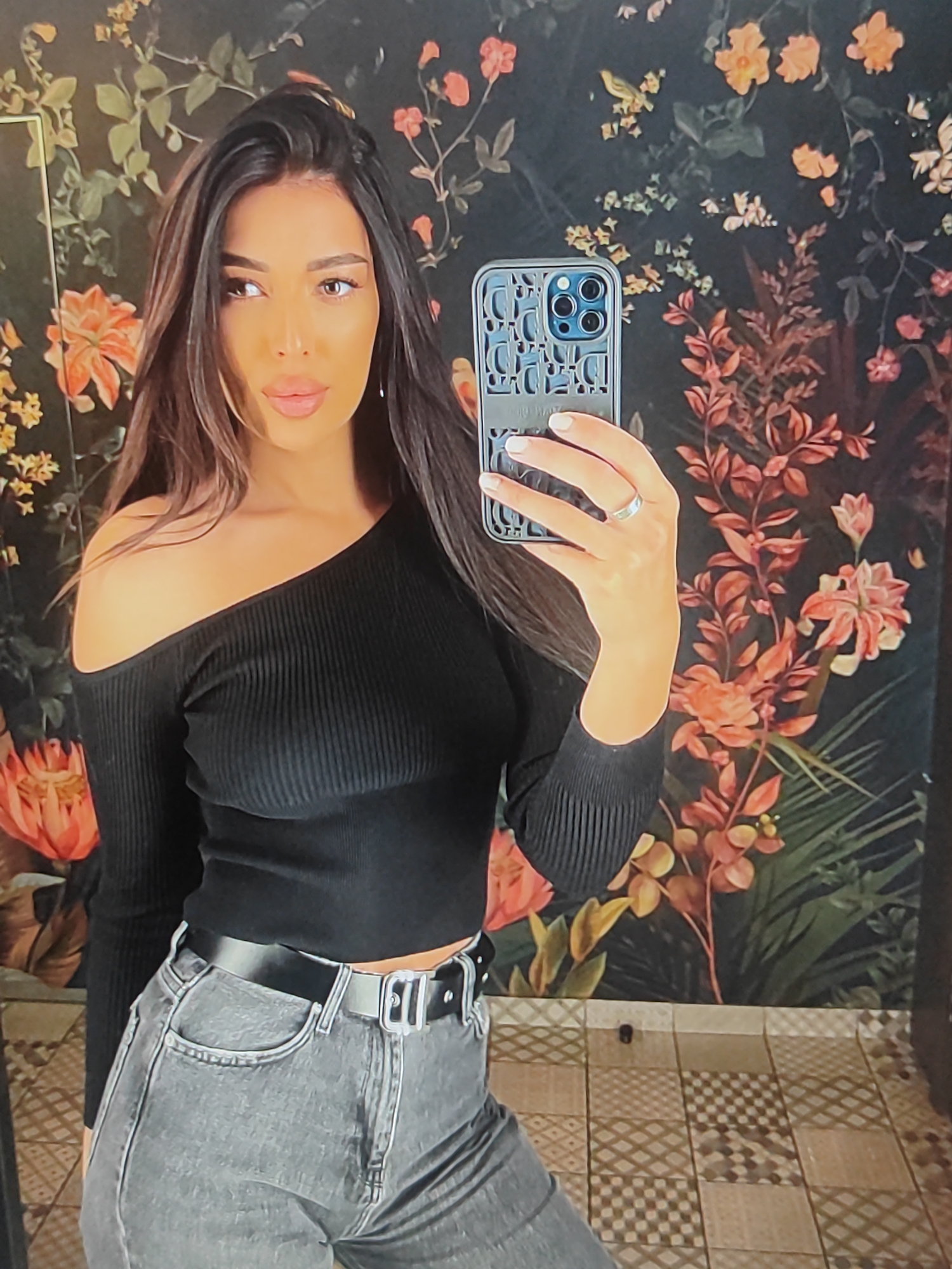 This is a medium shot, selfie-style photo of a woman with long, dark brown hair. She is wearing a black, off-the-shoulder, ribbed crop top and grey high-waisted jeans with a black belt. She is holding a smartphone in her right hand, which has a distinctive patterned case. A ring is visible on her left ring finger. The woman is looking at the camera with a neutral expression.
The background features a dark, floral wallpaper with large, stylized flowers in shades of orange, yellow, and pink, interspersed with green leaves. The floor is tiled with a mosaic pattern of square tiles in earthy tones, creating a diamond-like effect. The lighting suggests an indoor setting, possibly a restroom or a dressing room, with soft illumination. There is no indication of the time of day or weather conditions. The overall impression is a posed, casual portrait.
The phone case has a repeating pattern of the Dior logo.