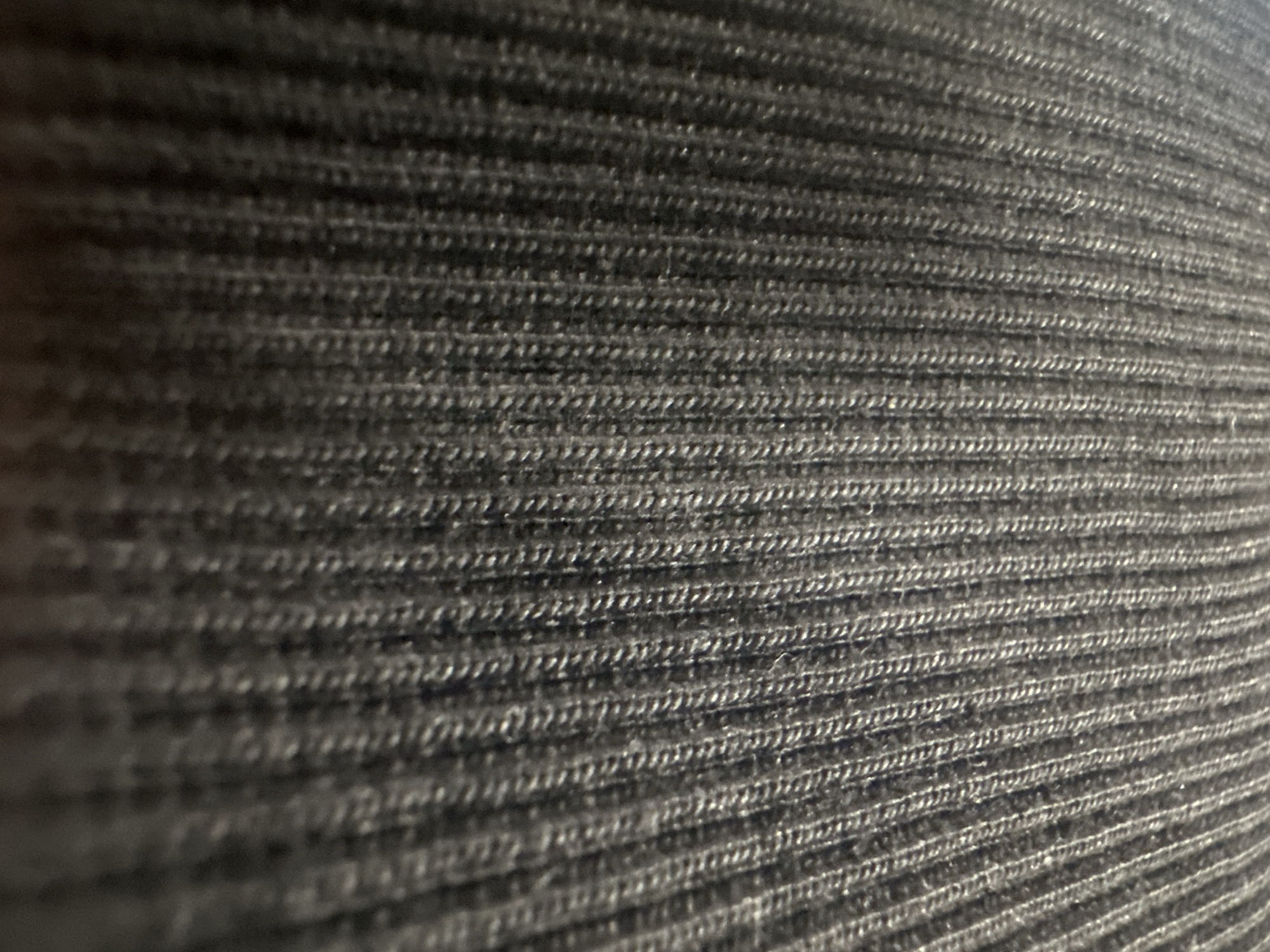 The image is a close-up, high-angle shot of a dark grey, ribbed knit fabric. The texture of the fabric, characterized by parallel vertical ridges, is the dominant feature. The lighting is uneven, creating shadows that accentuate the ribbed pattern. The background is also this fabric. There is no discernible location within the city of Letterkenny, Ireland, visible in this image, as it is a close-up of a textile.