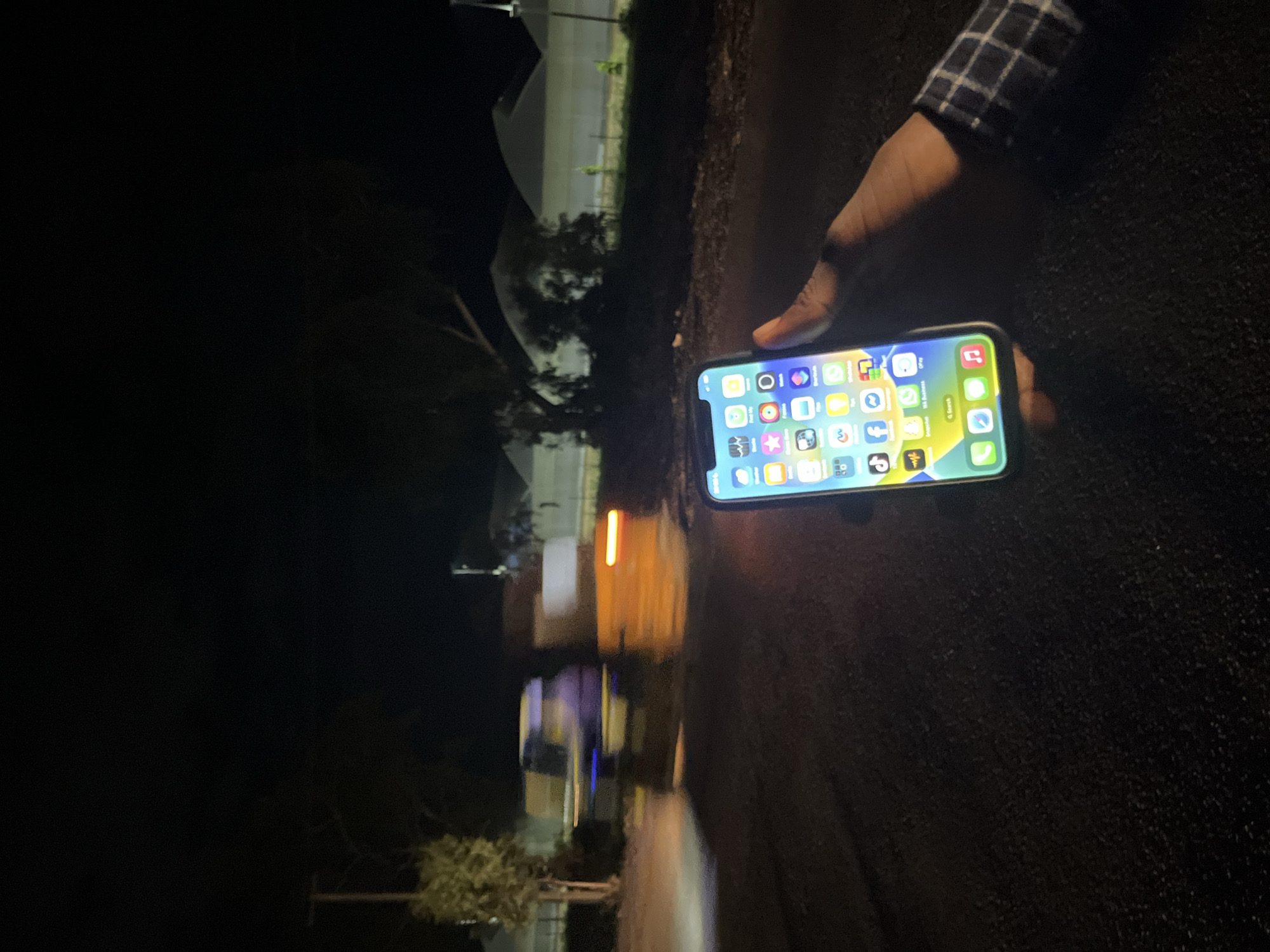 The image depicts a nighttime outdoor scene on an asphalt road in Jalingo, Nigeria. In the immediate foreground on the right, a dark-skinned right hand, partially covered by the cuff of a blue and white checkered shirt, holds an illuminated iPhone. The iPhone's screen displays its home interface, populated with multiple application icons arranged in rows, with the time "20:45" visible in the top-left corner of the status bar. In the mid-ground, two vehicles are in motion, rendered with significant blur. The vehicle on the left appears primarily blue/purple with visible headlights, while the following vehicle on the right is yellow/orange and exhibits a distinct red rear light. In the background, dark trees and bushes are visible on either side of the road, along with a utility pole on the left. Further in the distance, several large, white, arched structures, consistent with greenhouses or similar agricultural shelters, are discernible and appear to be under artificial illumination. The overall scene is dimly lit, with primary light sources emanating from the smartphone screen, the vehicles' lights, and the distant structures.