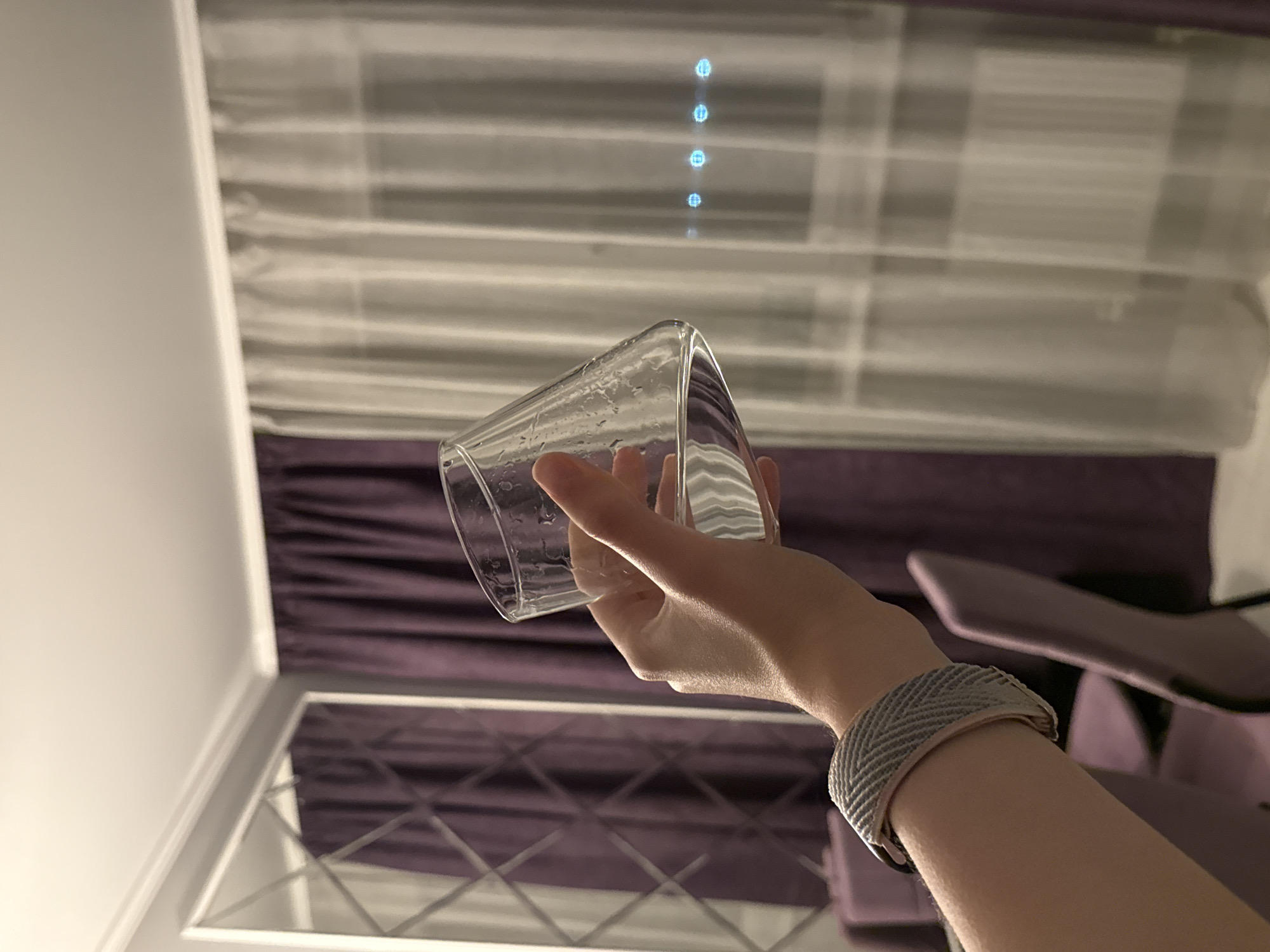 The image shows an indoor scene, appearing to be rotated 90 degrees counter-clockwise from its natural orientation. In the foreground, a person's bare hand and forearm are visible, holding a clear glass tilted downwards. The glass contains numerous water droplets or condensation, suggesting it recently held a cold liquid or is affected by humidity. A woven wristband, possibly a watch strap, in shades of grey and light pink, is worn on the person's wrist.
The background reveals parts of a room. Prominently featured are purple curtains or drapes that cover a significant portion of the wall. Behind or through the slats of what appears to be blinds, several distinct, vertically aligned blue-white light sources or reflections are visible. Below the main curtain section, a white wall is seen. Further down, a reflective surface, likely a mirror or a glossy table, displays a geometric crosshatch pattern and reflects the purple curtains above. To the far right, a piece of purple upholstered furniture, possibly a chair or headboard, is partially visible.
The lighting indicates an indoor setting, likely in the evening or at night, illuminated by artificial light. There is no visible text in the image.