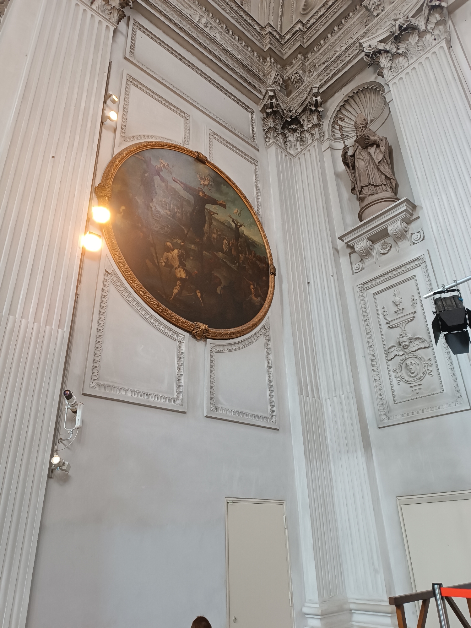 The image shows an interior view of a grand hall or church in Munich, Germany. The architecture is ornate, with tall fluted columns, decorative moldings, and intricate relief carvings on the walls. Dominating the left side of the frame is a large, oval painting in a golden frame, depicting a scene with several figures. To the right of the painting, a statue of a robed figure, possibly a saint or religious leader, stands in a niche. There are several lights and a projector visible in the scene, suggesting the space might be used for events or performances. The walls are painted a neutral, light color, and two doors are visible on the right side of the image. The overall impression is one of historical and architectural grandeur.