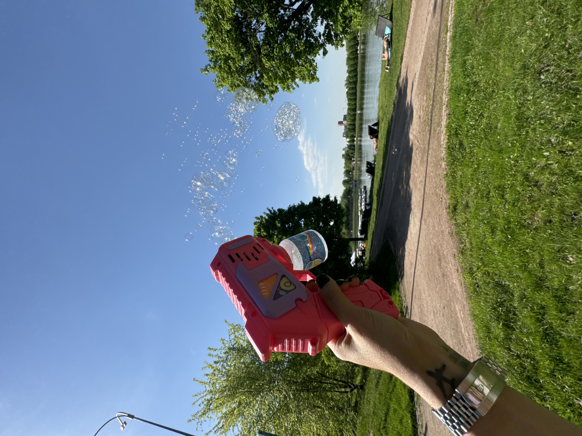 A person is holding a pink plastic bubble gun that is shooting bubbles into a clear blue sky. The bubbles are reflecting the sunlight. In the background, there is a body of water lined with green grass and trees. Some people are relaxing on the grass. The location is Gorky Park in Moscow, Russia. The time of day is daytime, likely midday, judging by the light.