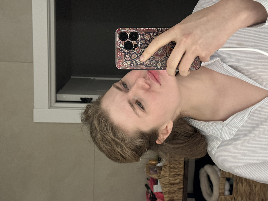 A person takes a selfie using a phone with a patterned case. The person has fair skin, light brown hair, and is wearing a striped button-down shirt. The background includes a bathroom vanity with a closed cabinet, beige walls, and a glimpse of a wicker basket with contents. The angle is slightly tilted.