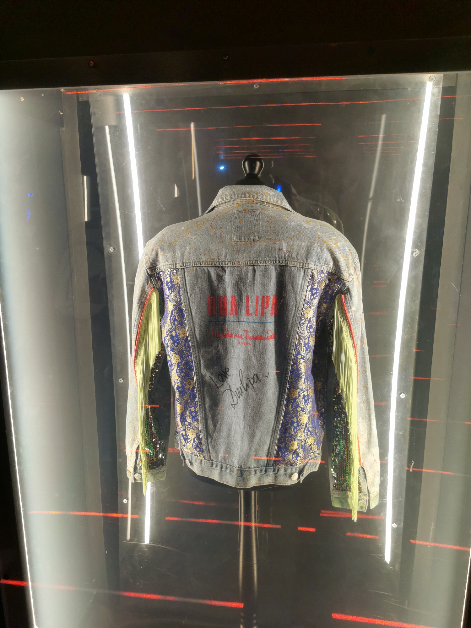 A denim jacket displayed in a glass case. The jacket features the text "Dua Lipa" in red letters, with the text "Future Nostalgia" in smaller font underneath. The jacket is decorated with blue and gold floral patterns and has green and gold fringe on the sleeves. The signature "Love, Dua Lipa" is visible on the lower back of the jacket. The case is illuminated by vertical white lights, and horizontal red lights are reflected on the inside. The location is likely in London, United Kingdom.