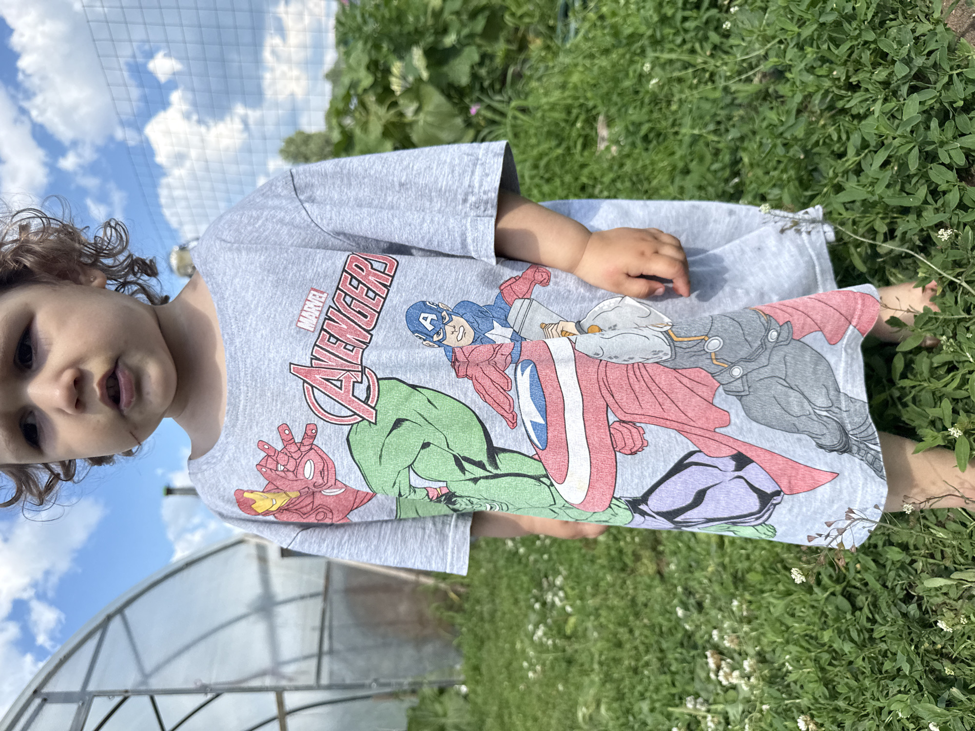 A young child stands in a grassy outdoor area, facing forward, with a greenhouse structure and vegetation in the background. The child is wearing a light gray t-shirt adorned with a graphic print of several Marvel Avengers characters, including Iron Man, Hulk, Captain America, and Thor. The t-shirt extends down to the child's mid-thigh. The background features lush green grass, small white flowers, and the curved, transparent structure of a greenhouse. The sky above is blue with scattered white clouds. The scene suggests a casual, possibly domestic setting, with the child being the primary subject of the image.