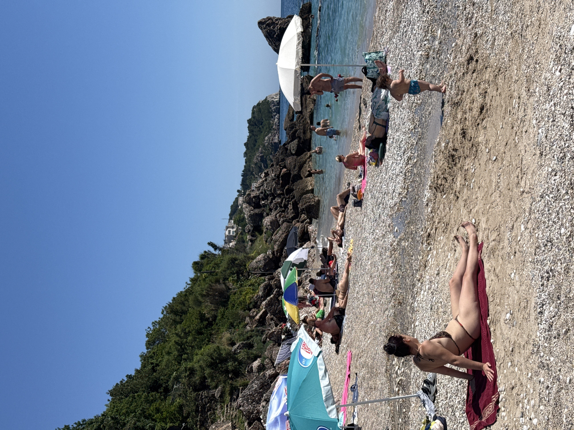 This detailed image depicts a bustling pebble and sand beach in Drobnići, Montenegro, under a clear, bright blue sky. The scene is illuminated by strong daylight, casting distinct shadows.
The beach itself consists primarily of small, light-colored pebbles, with a darker, damp sandy area visible in the foreground. To the left and in the background, steep, rocky cliffs covered in dense green vegetation ascend, with a few white buildings with red roofs discernable on the distant hillsides. The water is clear, calm, and turquoise-blue, reflecting the sky. Large, dark grey rock formations punctuate the coastline, both on the beach and partially submerged in the water, creating natural coves and features.
Approximately 20-25 individuals are visible on the beach and in the water. In the immediate foreground, a woman with dark hair tied in a bun is sunbathing on a red towel, lying on her back with her head turned to the right. She is wearing a leopard-print string bikini. To her right, a small child, a toddler, in blue patterned swim trunks, stands barefoot on the sandy part of the beach, looking to the right.
Further into the mid-ground, numerous people are engaged in beach activities. Several individuals are lying on towels and mats of various colors (pink, green, patterned), sunbathing or resting under the shade of multiple beach umbrellas. Visible umbrellas include a large turquoise one with a partial "Nestle" logo on the left, a blue and white striped umbrella, a yellow, blue, and green umbrella, and a prominent white umbrella on the right.
In the water, several adults are swimming or wading. One man stands under the white umbrella, looking at a device in his hand. Another man stands in the shallow water, facing away from the camera. The overall atmosphere suggests a leisurely summer day at the coast.
