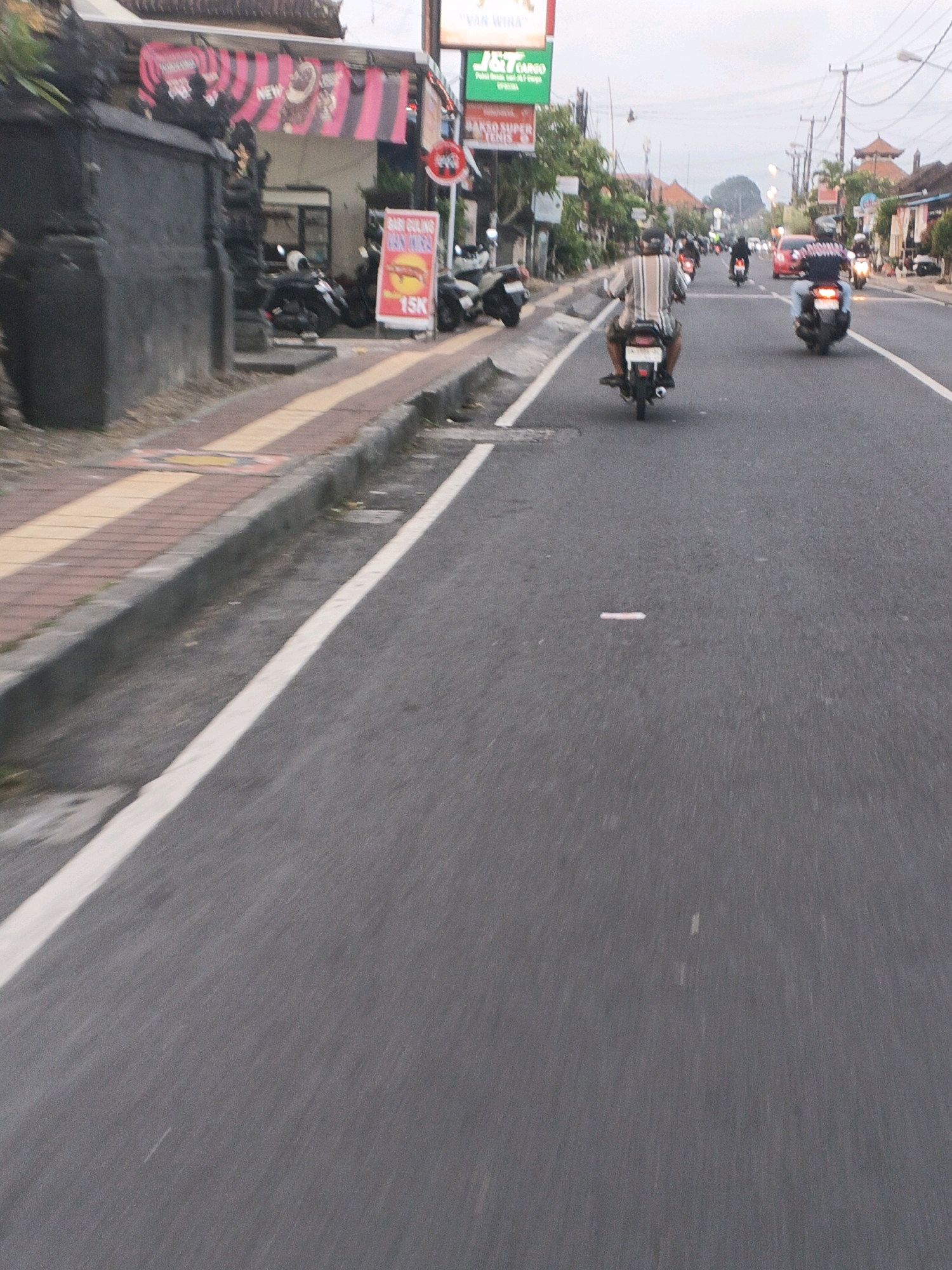 The image captures a street scene in Denpasar, Indonesia, likely during daytime based on the lighting. The foreground is dominated by a paved road with a white dashed line marking the center. The perspective suggests the photo was taken from a moving vehicle or motorcycle. On the left side of the road, there is a concrete curb and a sidewalk with patterned paving stones. Adjacent to the sidewalk is a dark, textured wall and a building facade with pink and black striped banners and signs. Several parked motorcycles are visible in front of the building.
Further down the street, traffic is moving. A man on a motorcycle, wearing a striped shirt, is in the immediate middle ground. Behind him, other motorcycles and at least one red car are also present, indicating moderate vehicular activity. The background shows more buildings, trees, and utility poles against a slightly overcast sky. The overall impression is of a typical urban street in Indonesia, characterized by the prevalence of motorcycles and local businesses.
