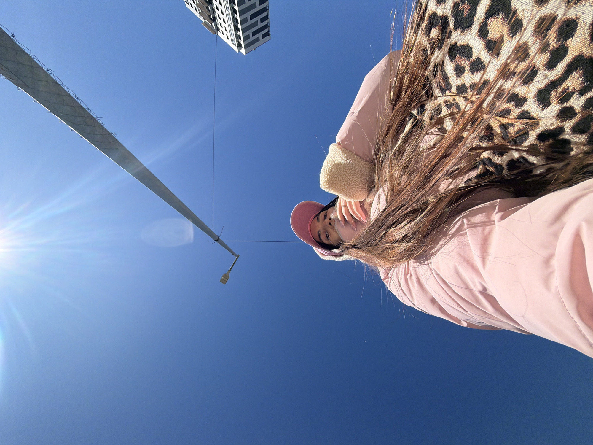 The image presents a low-angle perspective of a woman wearing a pink jacket with a fur collar, and a pink baseball cap. She is also wearing a leopard print scarf or wrap. Above her, there is a clear blue sky, a tall building, and a light pole, possibly a street lamp, visible. The location could be Saint Petersburg, Russia, based on the architectural style of the building.
