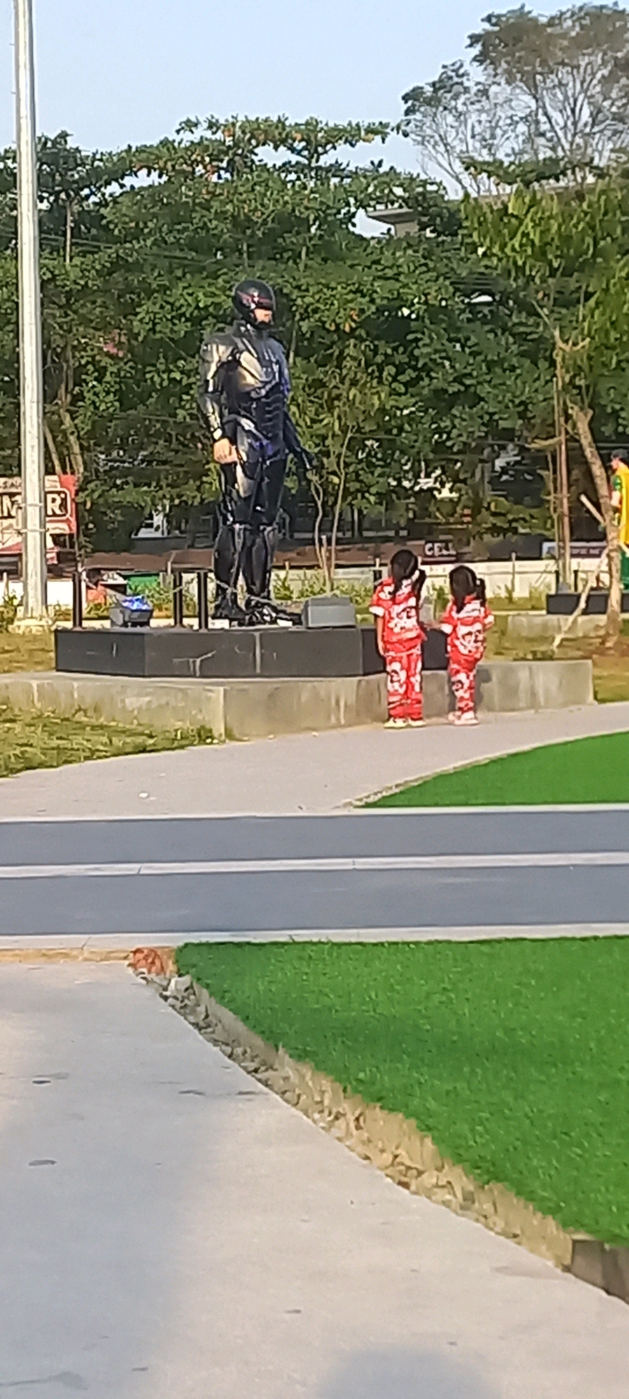 An outdoor statue of RoboCop stands on a raised platform. Two young girls in red and white patterned outfits stand to the right of the statue, facing away from the camera. The statue is dark and metallic, with visible details of its armor and helmet. The girls have dark hair. The background includes lush green trees and a clear sky. A street with two white lines runs horizontally in the foreground, separating a paved walkway from a patch of green grass. A curb with rocks is visible between the walkway and the grass. A white pole stands to the far left of the image.