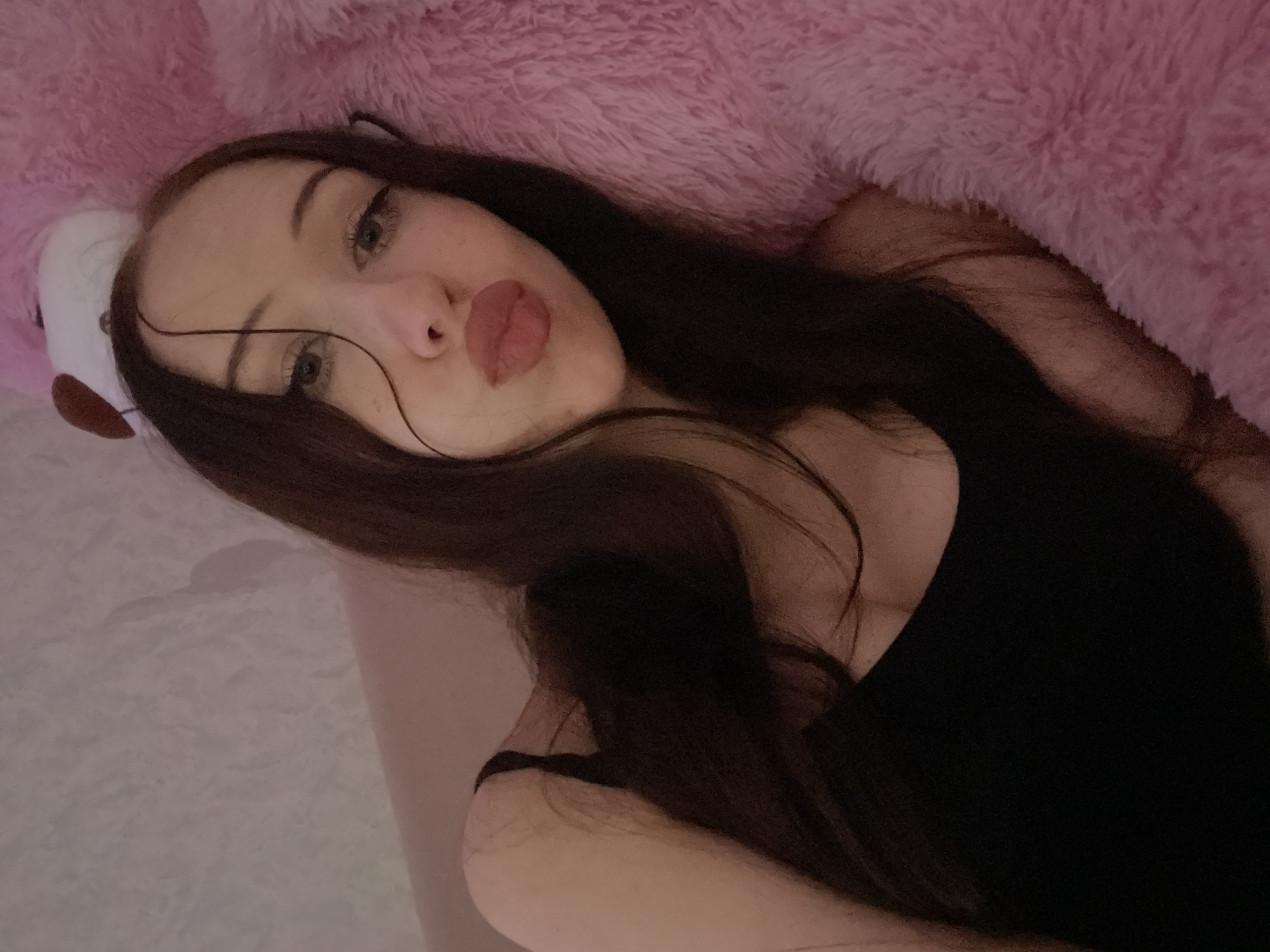 The image shows a young woman with long, dark hair, wearing a black tank top. She is lying down, possibly on a bed, with a large pink furry object to her right. Her face is turned towards the camera, and she is making a duck face. The background appears to be a textured wall in a soft pinkish hue. There are no discernible elements in the image that can be used to determine the specific location within Votkinsk, Russia.